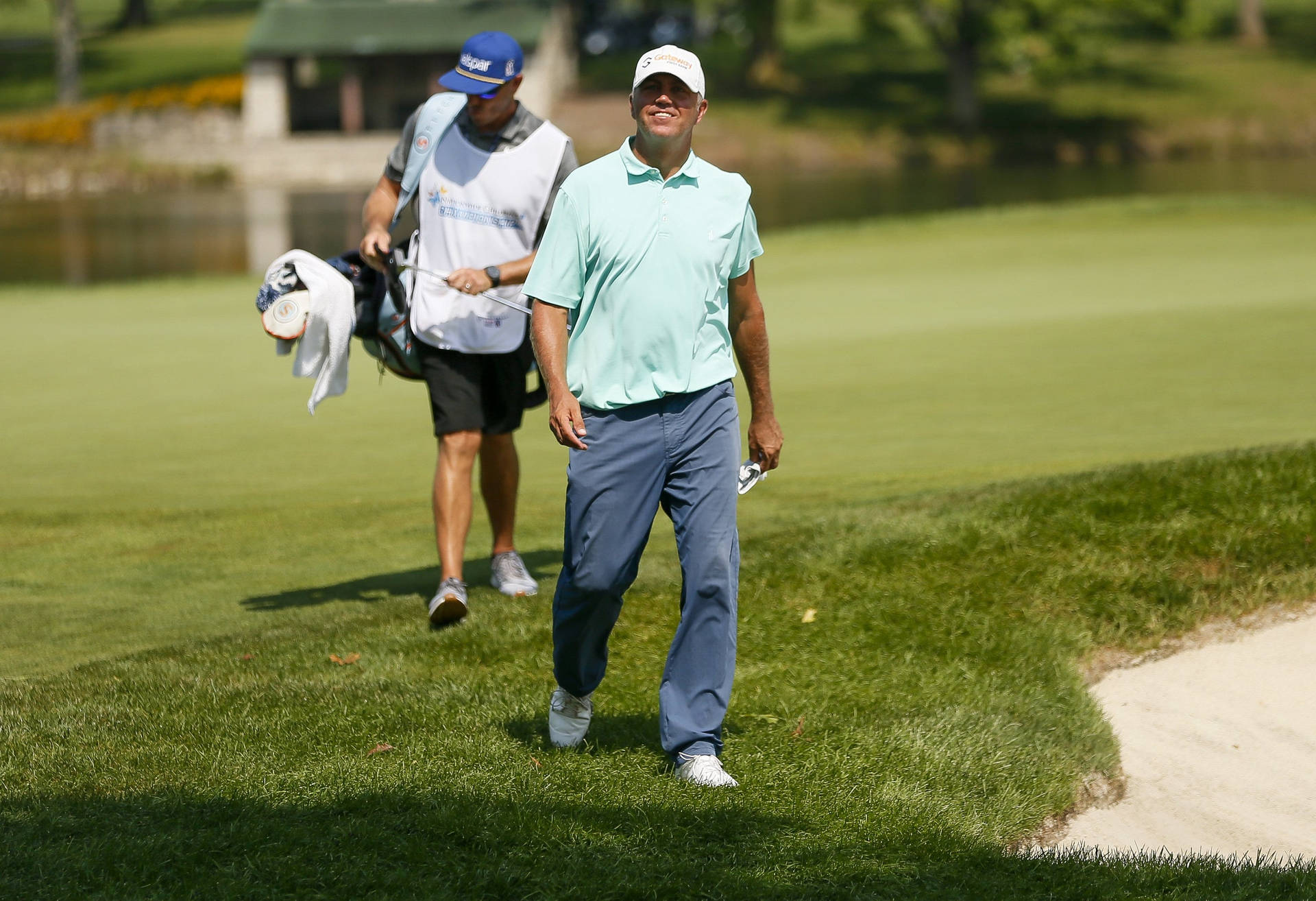 比爾·哈斯自信邁步於高爾夫球場 壁紙 — 一位身穿淺藍色襯衫、面帶微笑的高爾夫球手自信地走過陽光明媚的綠色高爾夫球場，他的球童跟在後面。