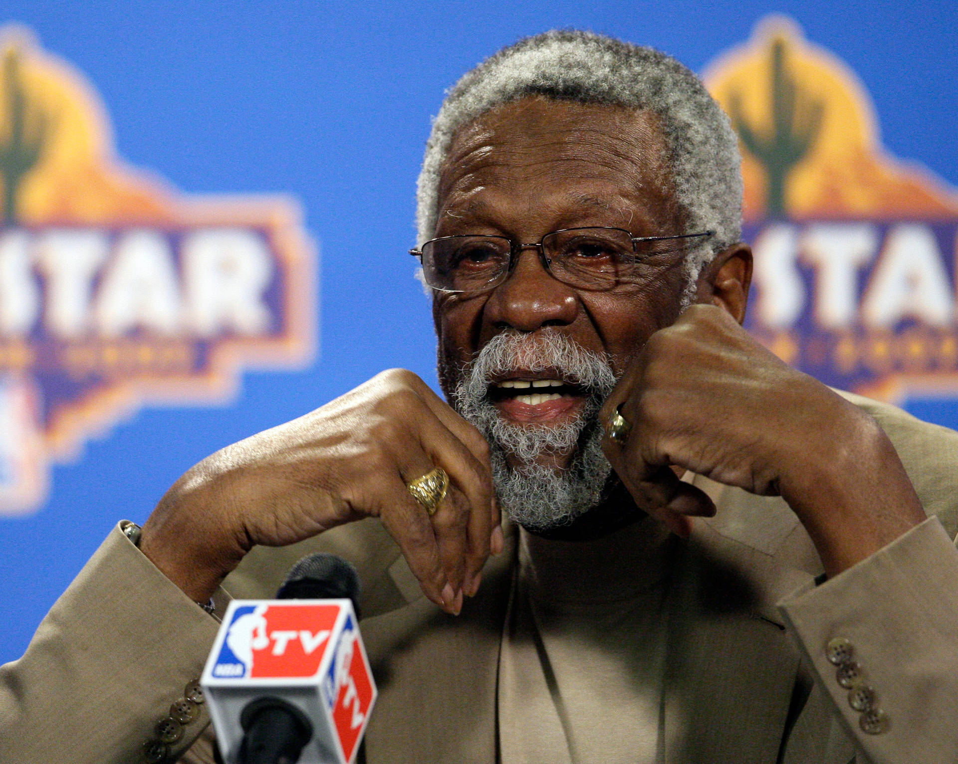 一位戴眼鏡、留灰鬍子的年長黑人男子，在設有NBA TV咪高峰的新聞發布會上微笑。