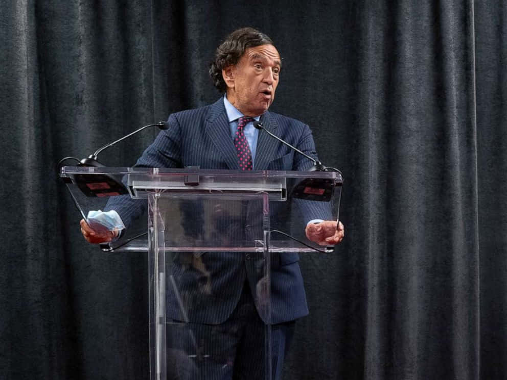 穿西裝的男子在講台發言，手持口罩。