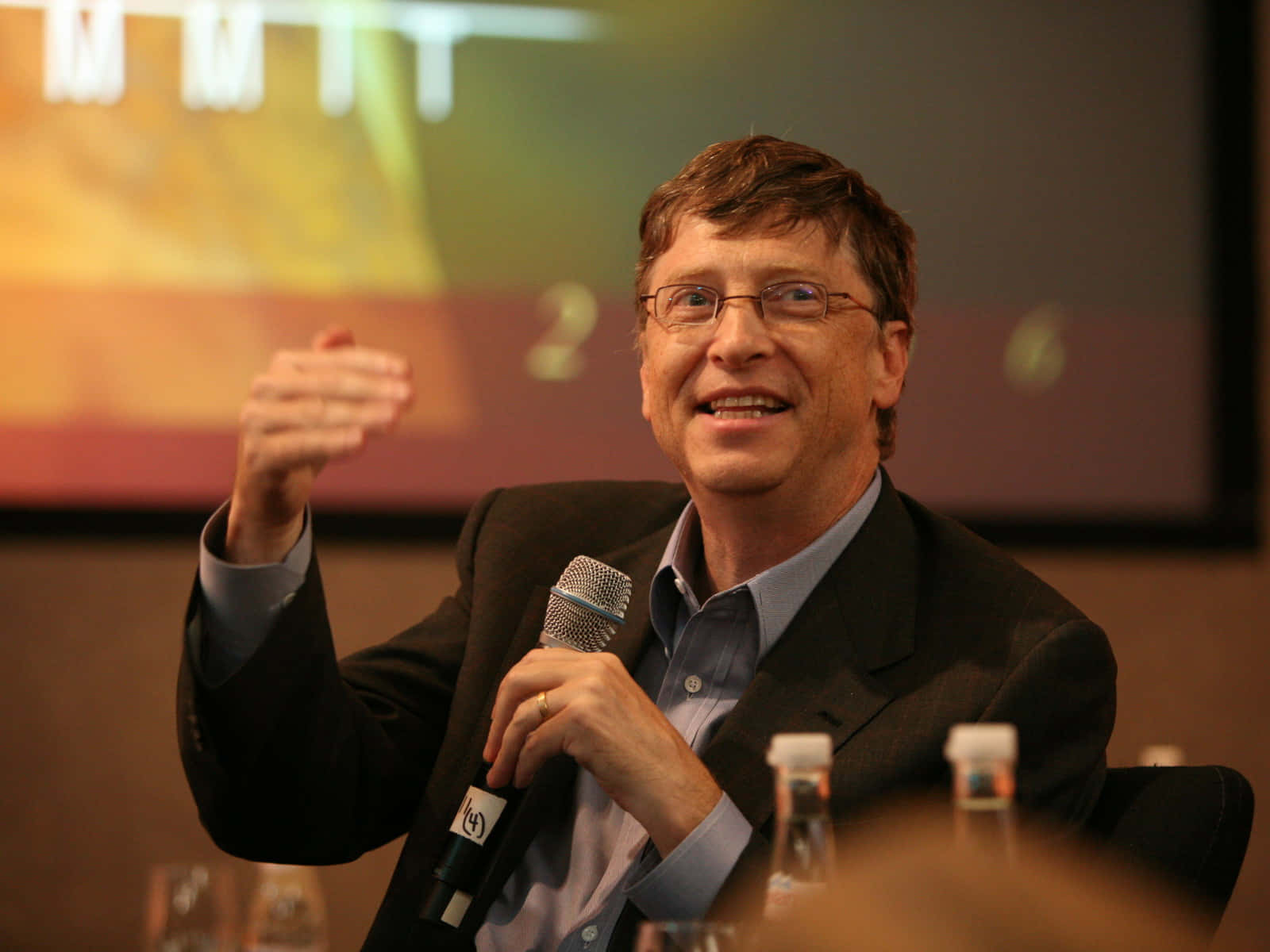 Bill Gates 桌布 壁紙 — 比爾·蓋茨在會議上對著麥克風溫暖地微笑說話，身後屏幕上可見「MIT」字樣。
