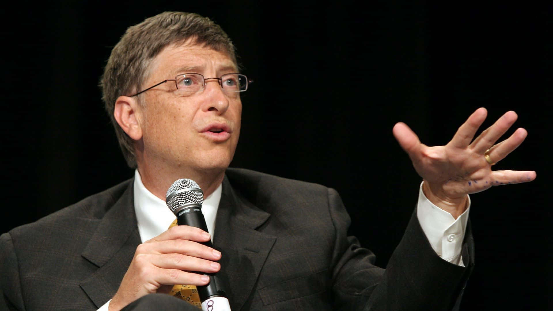 Bill Gates 桌布 壁紙 — 比爾·蓋茨在深色背景前用左手做手勢對著麥克風講話。
