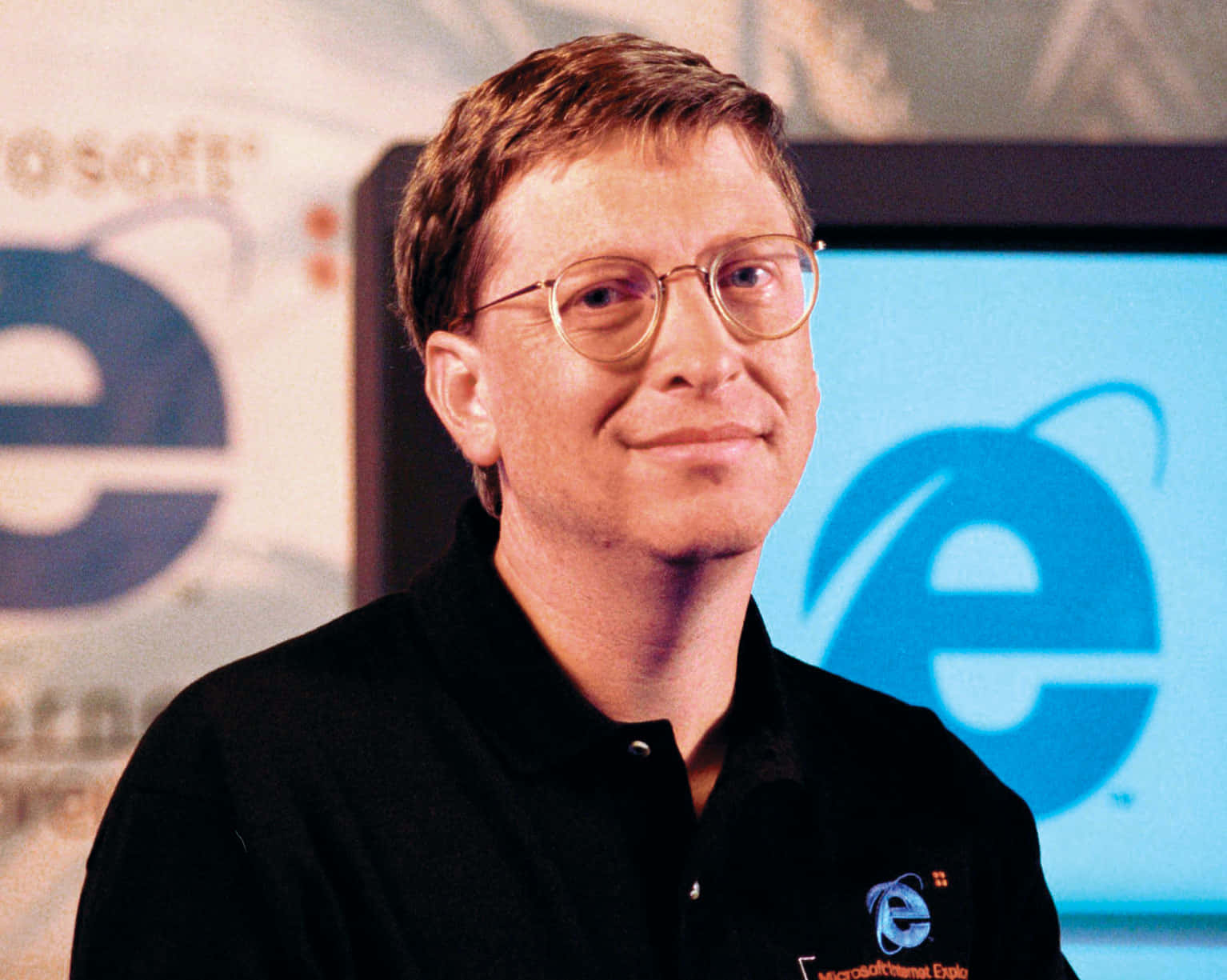 Bill Gates 桌布 壁紙 — 比爾·蓋茲穿著黑色Polo衫，站在螢幕上發光的藍色Internet Explorer標誌前微笑。