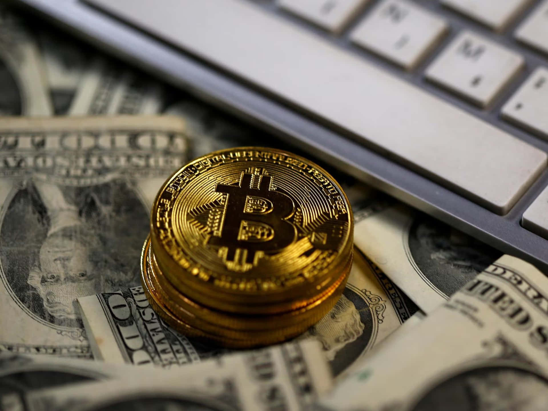 比特幣——金融交易的未來 壁紙 — 一堆閃亮的金色比特幣硬幣放在散落的美鈔上，旁邊是筆記本電腦鍵盤。