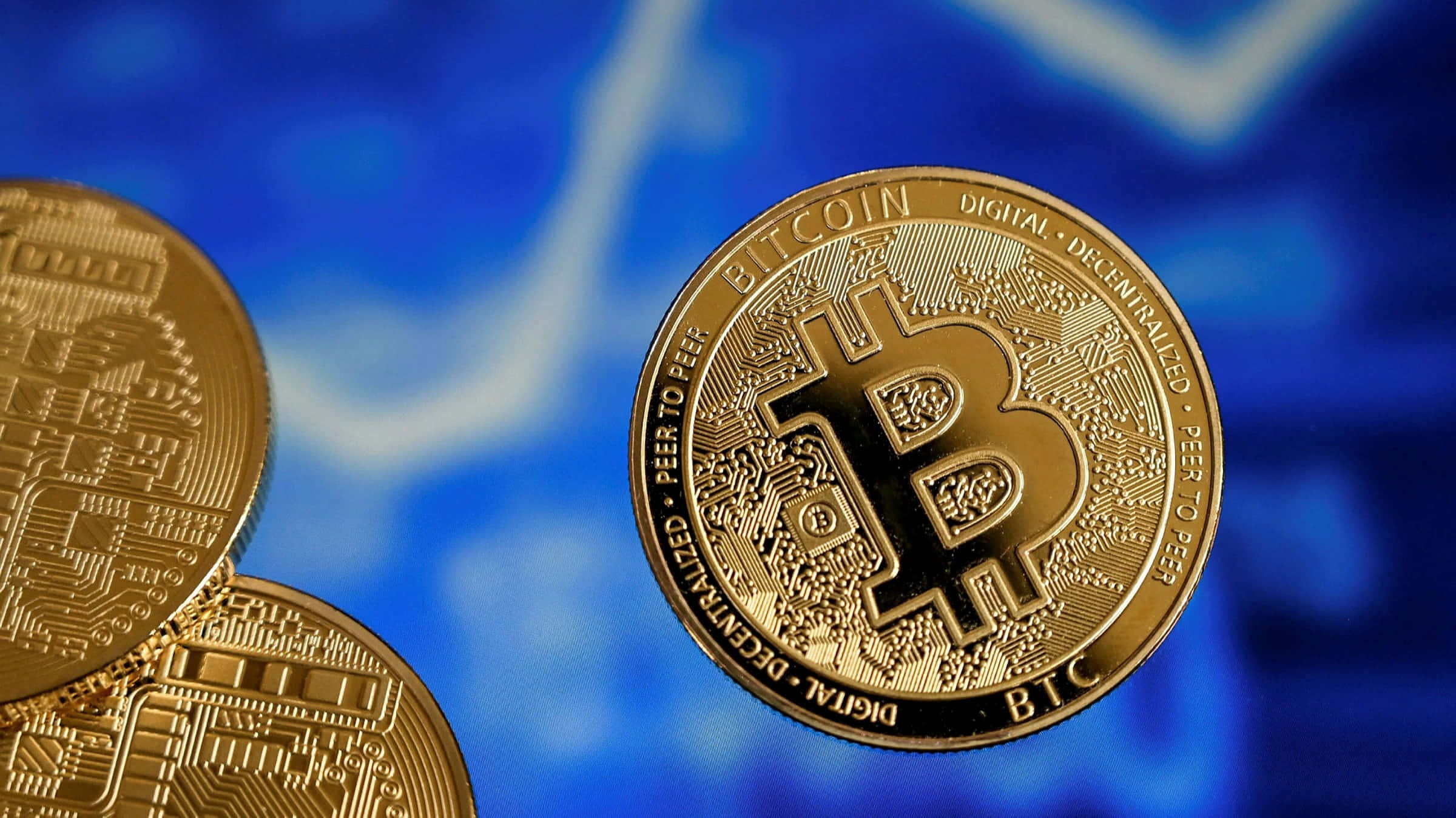 比特幣加密貨幣正改變我們對錢的思考方式 壁紙 — 金色比特幣硬幣在模糊的藍色金融圖表背景上清晰對焦。