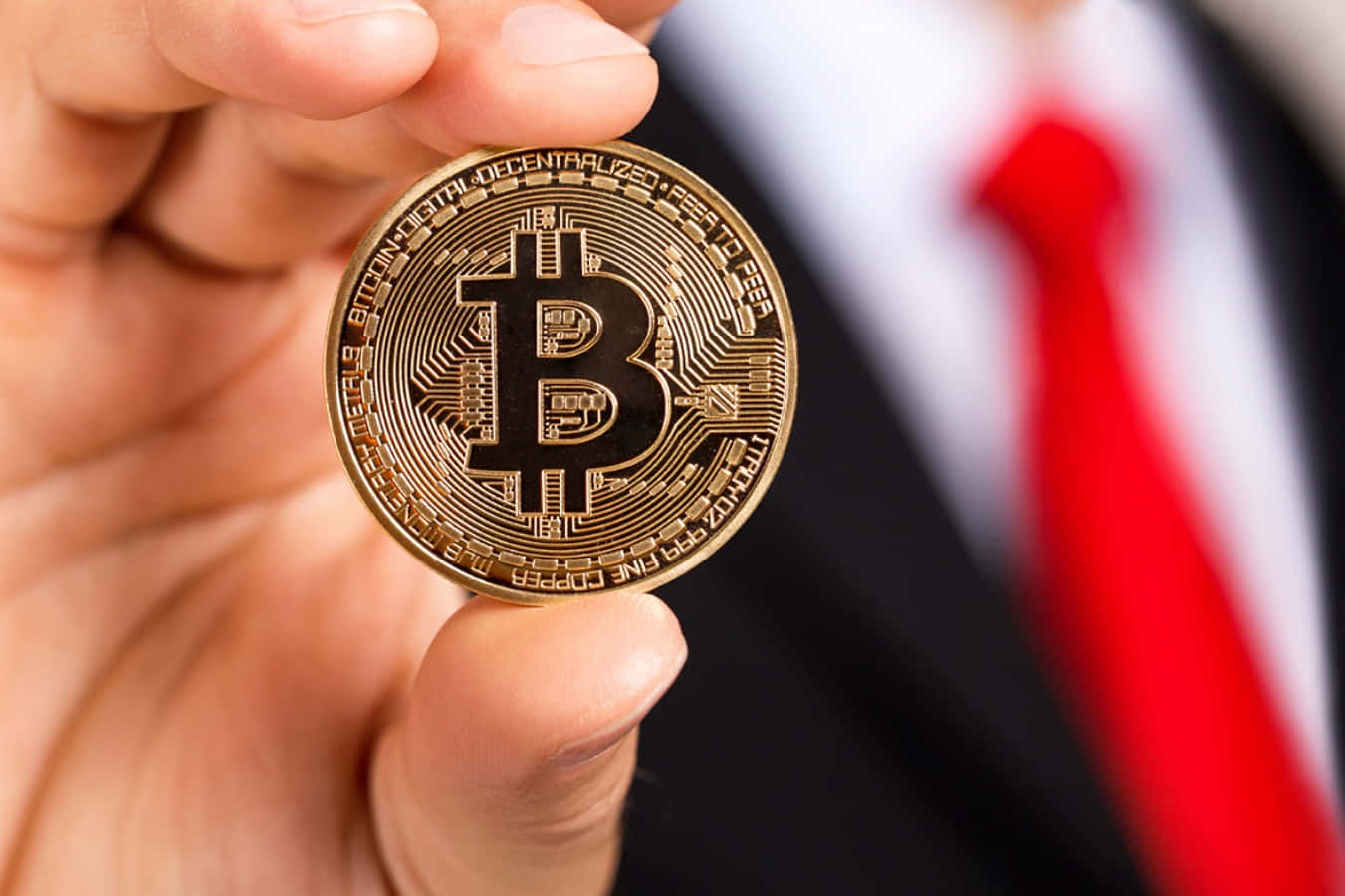 比特幣是數位貨幣的未來 壁紙 — 一隻手拿著帶有電路設計的金色比特幣硬幣，背景是模糊的穿著西裝和紅色領帶的商人。