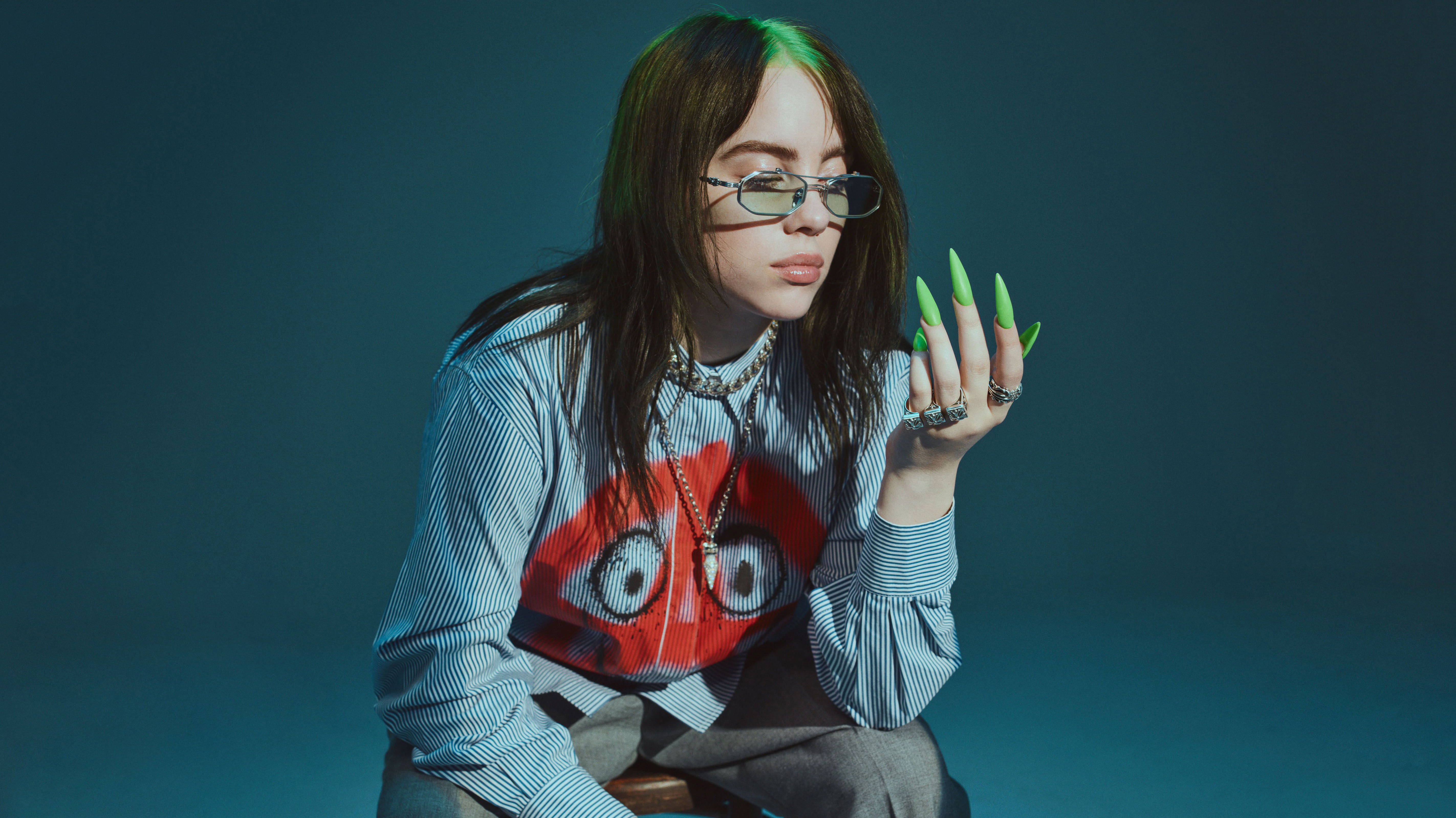 比莉·艾利什 4K 綠色指甲 壁紙 — Billie Eilish 戴著眼鏡，塗上螢光綠色指甲，身穿印有紅色卡通臉的條紋襯衫，在憂鬱的深藍綠色背景前擺姿勢。