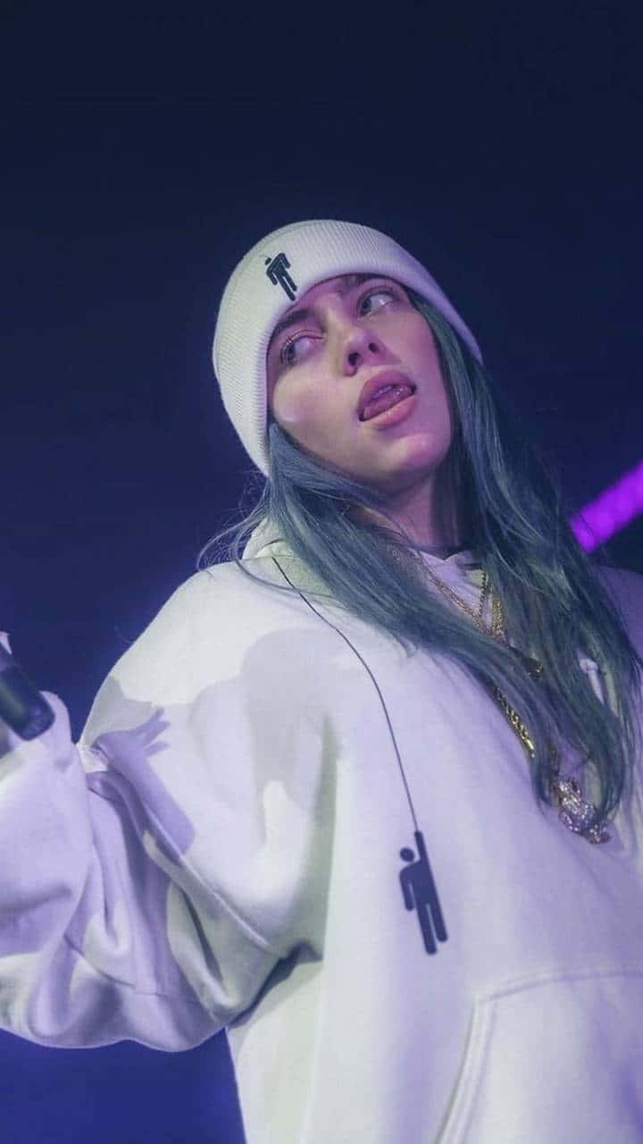 Billie Eilish 在紫色燈光下穿著白色連帽衫和冷帽在舞台上唱歌。