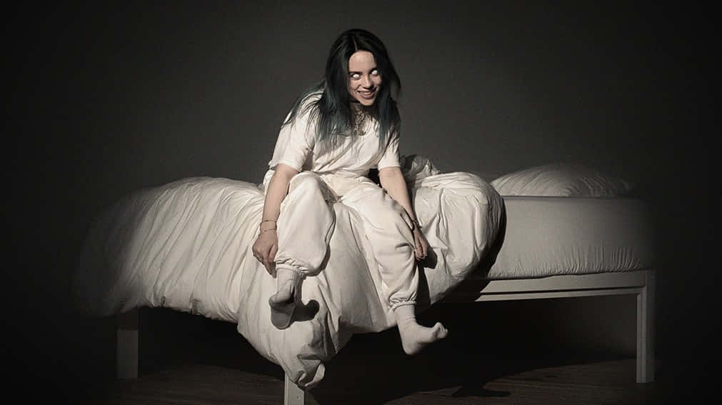 Billie Eilish 坐在黑暗房間的床上，身穿白色休閒服。