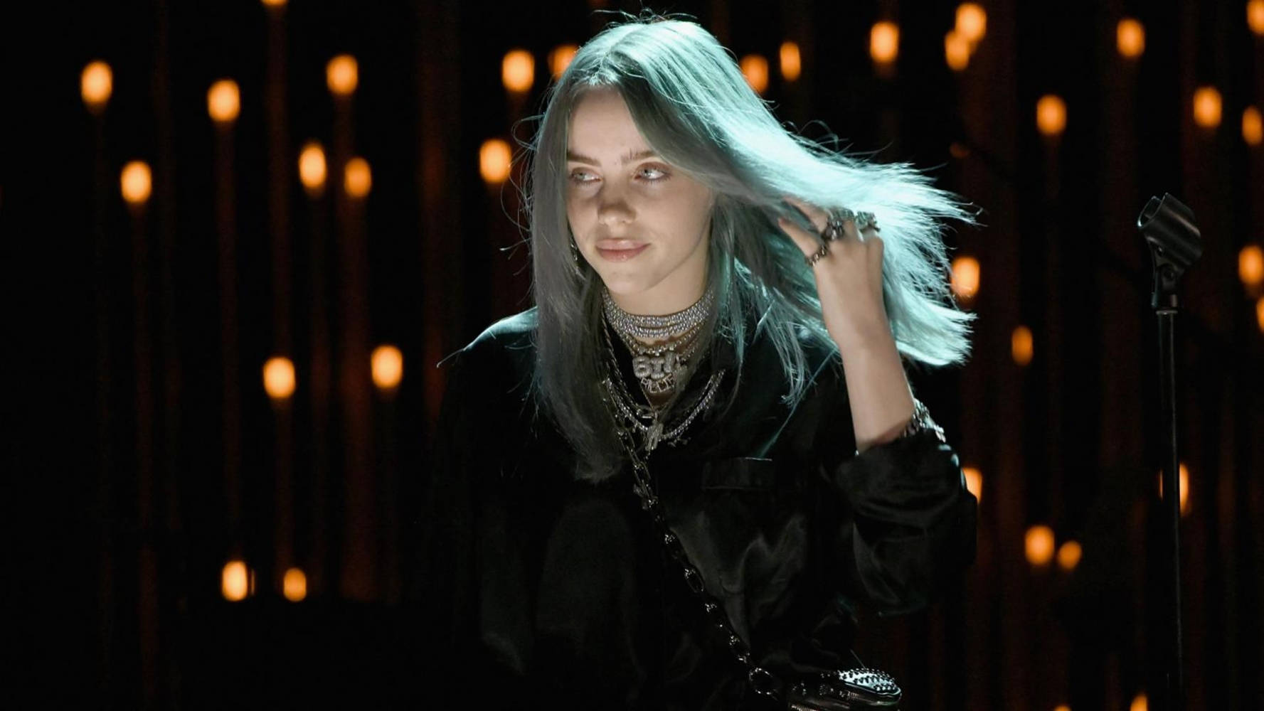 比莉·艾利什燈光特效美學照片 壁紙 — Billie Eilish 銀色頭髮和層疊項鍊站在舞台上，在溫暖的燈光下。