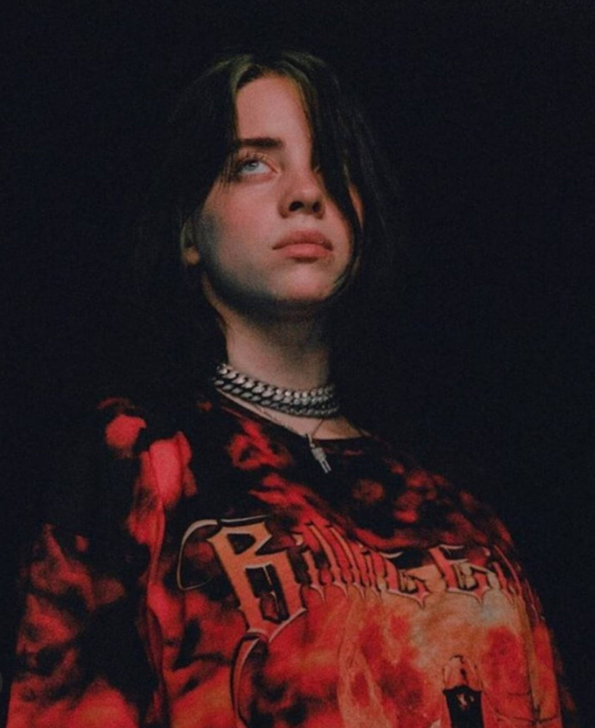 比莉·艾利什穿火焰美學T恤 壁紙 — Billie Eilish 在昏暗的肖像中向上凝視，身穿紅色圖案T恤和層疊銀鏈。