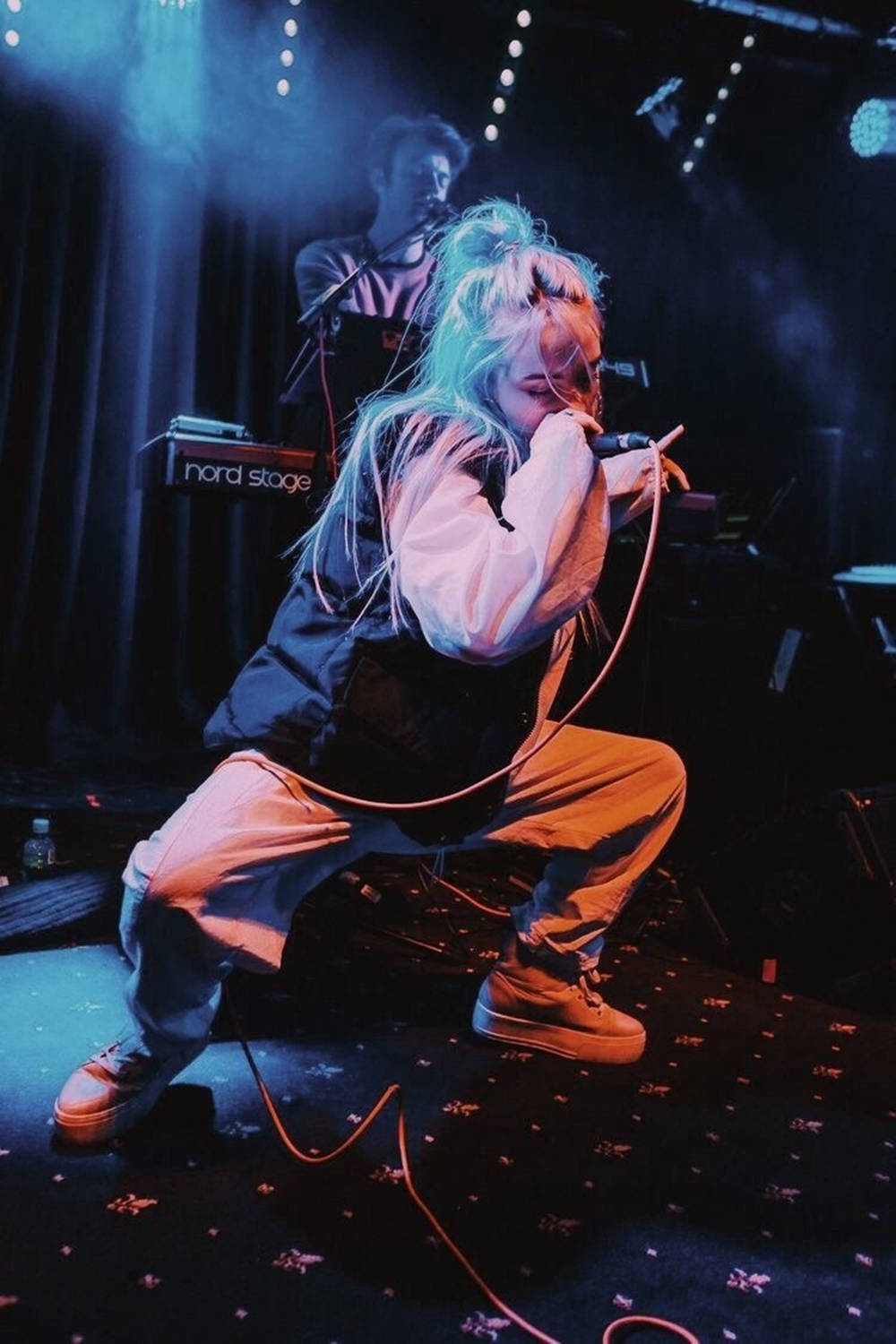 比莉·艾利什與樂隊演出美學壁紙 壁紙 — 一位擁有發光藍色頭髮的表演者蹲在舞台上，在戲劇性的藍色舞台燈光下對著麥克風唱歌。