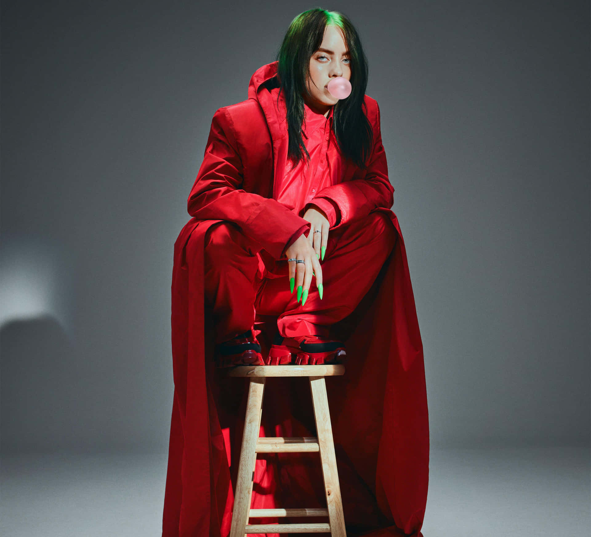 Billie Eilish 身穿紅色服裝在木凳上吹粉紅色泡泡糖泡泡。