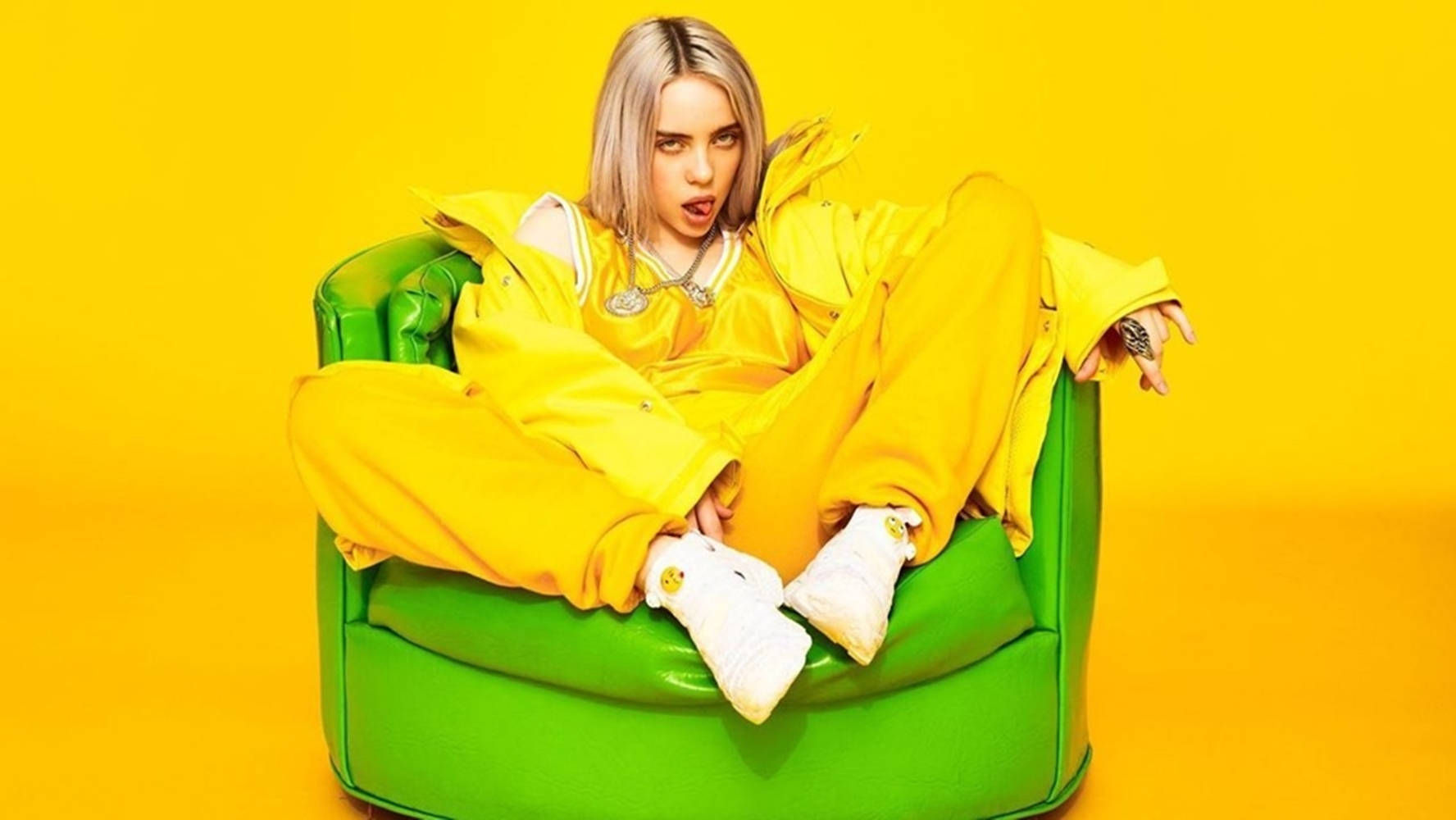 比莉·艾利什黃綠色美學壁紙 壁紙 — Billie Eilish 喺鮮黃色背景前，坐喺一張亮綠色嘅扶手椅上，着住一件大膽嘅黃色運動套裝。