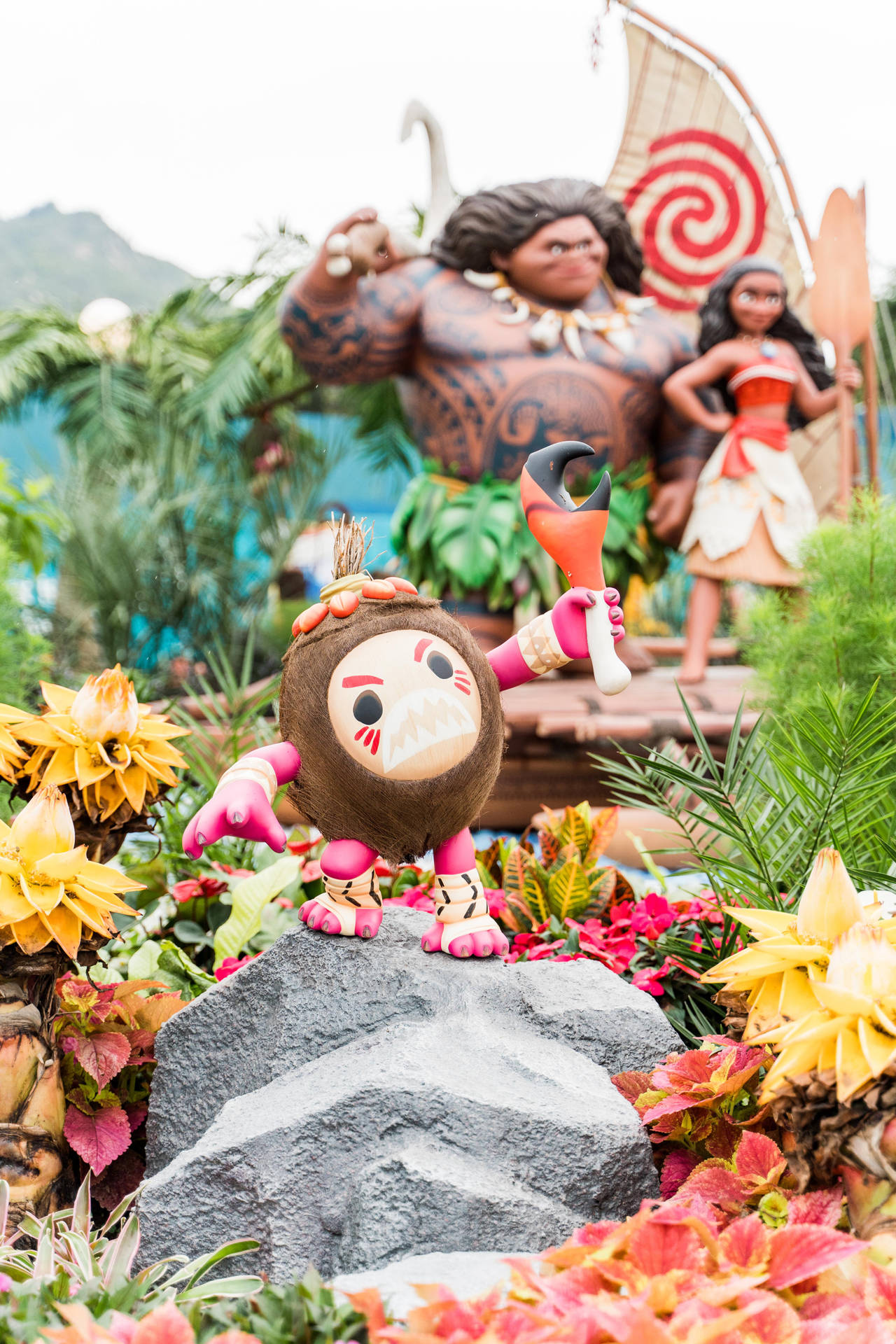 Maui 吉祥物被熱帶花朵包圍，後面有 Moana 和 Maui 雕像。
