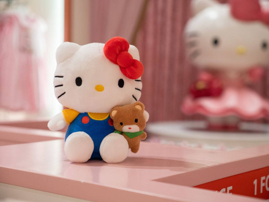 毛絨Hello Kitty卡通頭像 壁紙 — 一個穿著藍色工裝褲的毛絨Hello Kitty在粉紅色架子上抱著一個小泰迪熊，背景是模糊的粉紅色。