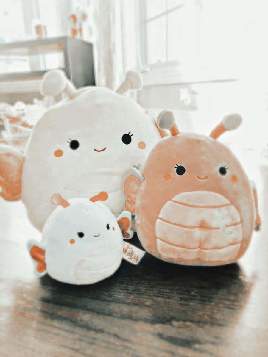 毛絨玩偶美學肖像背景 壁紙 — 三個可愛的毛絨Squishmallows，帶有柔和粉彩調，一起坐在靠近明亮窗戶的木質表面上。