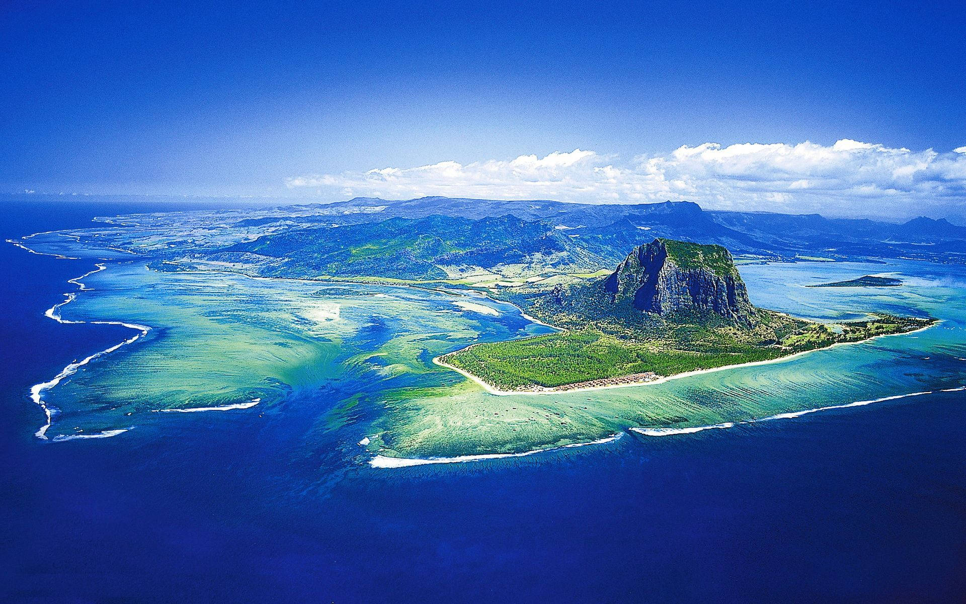 毛里求斯島 壁紙 — 從高空俯瞰一個鬱鬱蔥蔥的綠色島嶼，島上有一個突出的岩石山峰，周圍是充滿活力的碧綠海水和珊瑚礁，天空清澈湛藍。