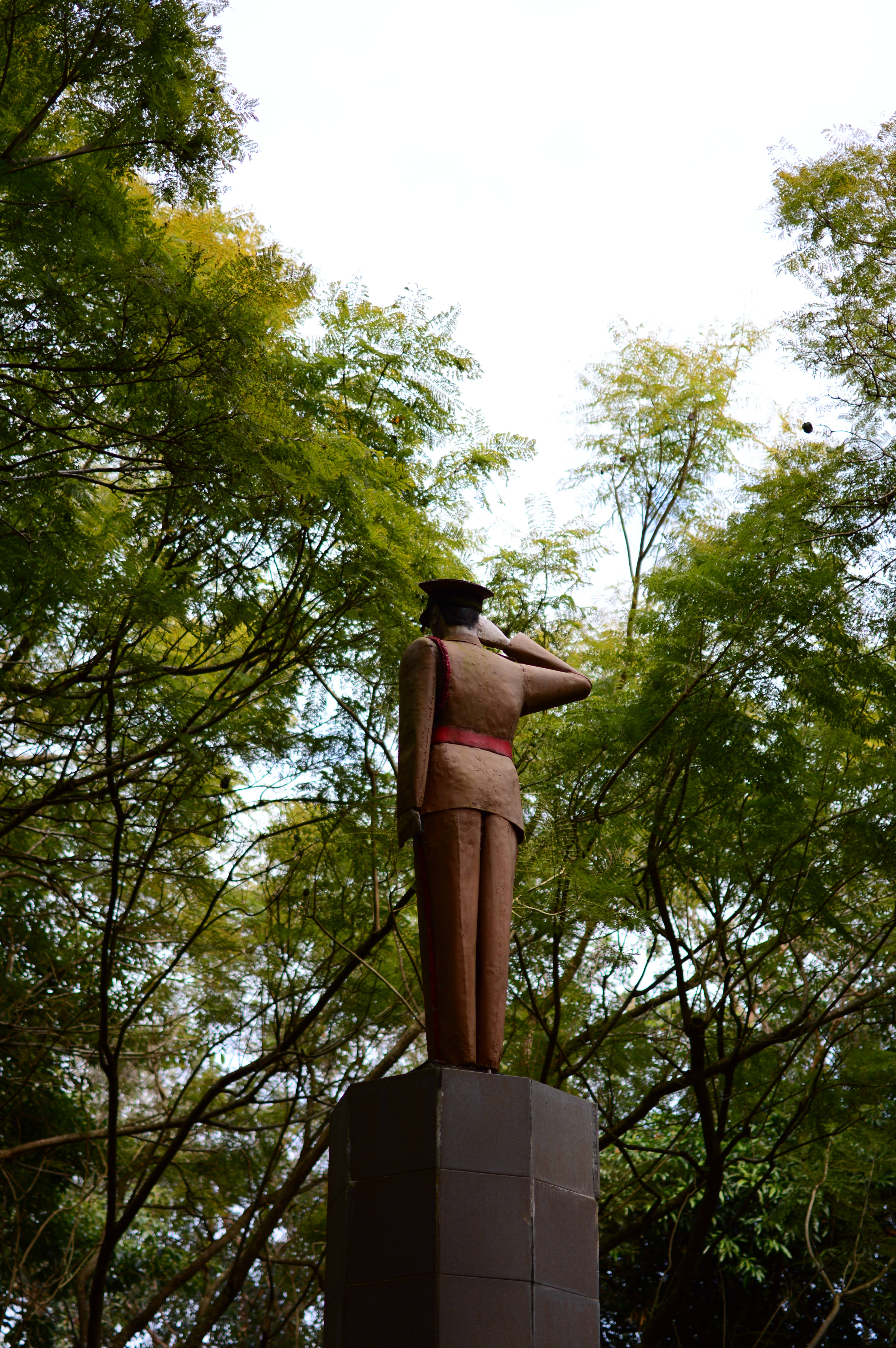毛里求斯島石雕 壁紙 — 一座青銅士兵雕像在基座上敬禮，周圍環繞著茂密的綠樹，天空明亮。