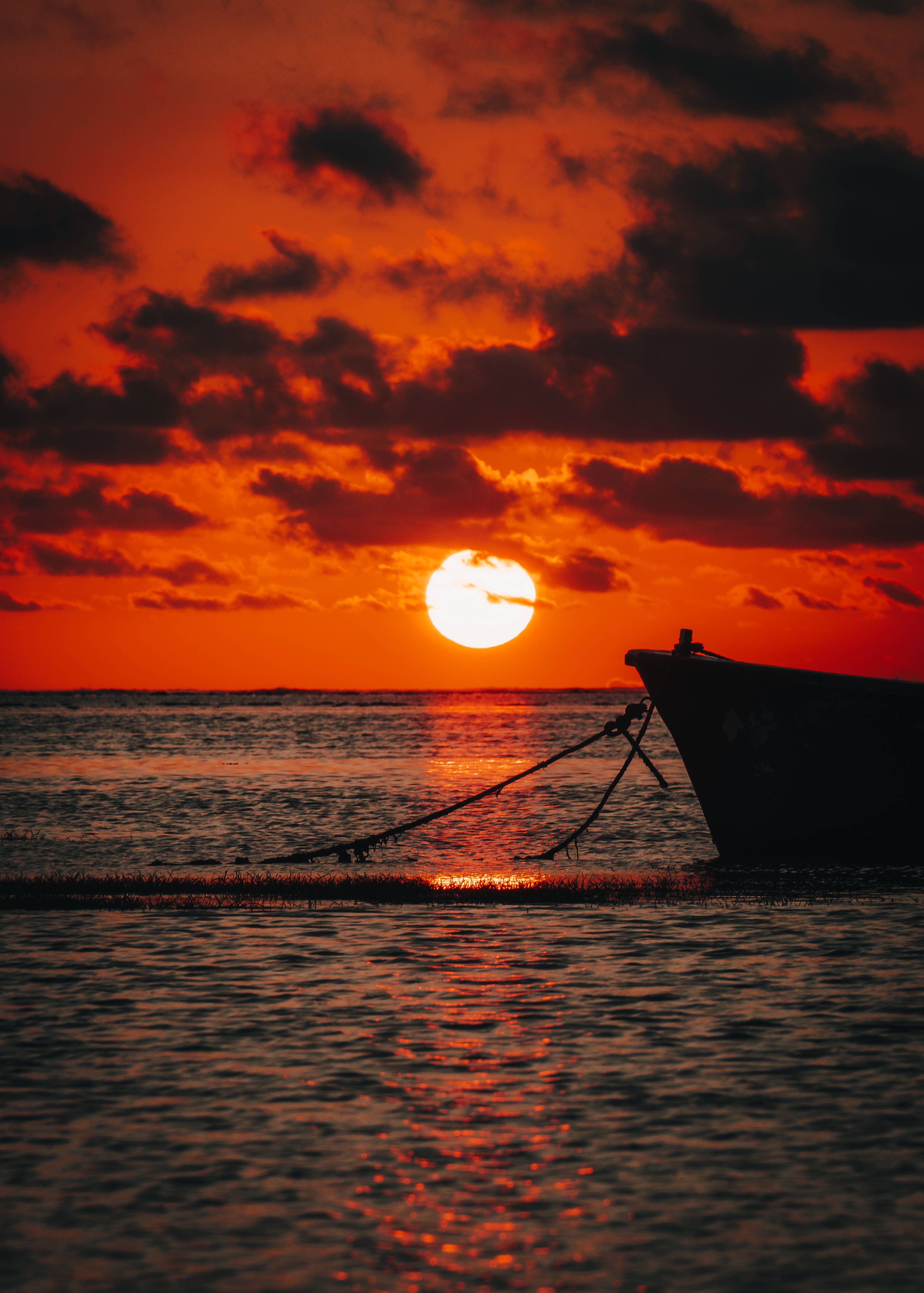 毛里求斯日落海船 壁紙 — 一艘漁船的剪影在發光的橙色水面上，太陽在戲劇性的雲層後落下。