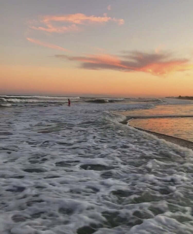 氣泡海灘美學圖片 壁紙 — 一個孤獨的身影在海洋波浪中涉水而行，粉紅色和橙色的雲彩在日落時分在地平線上發光。