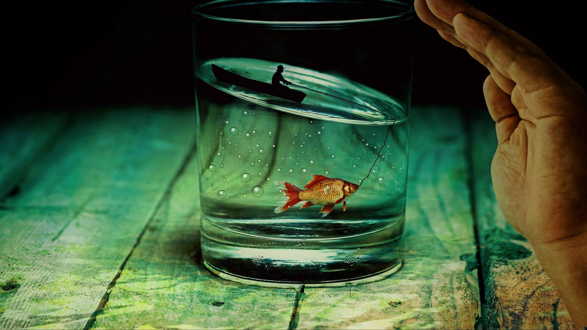一隻手拿著一個玻璃杯，裡面有一條金魚在釣魚線上，上方有一個微型小船和人物。