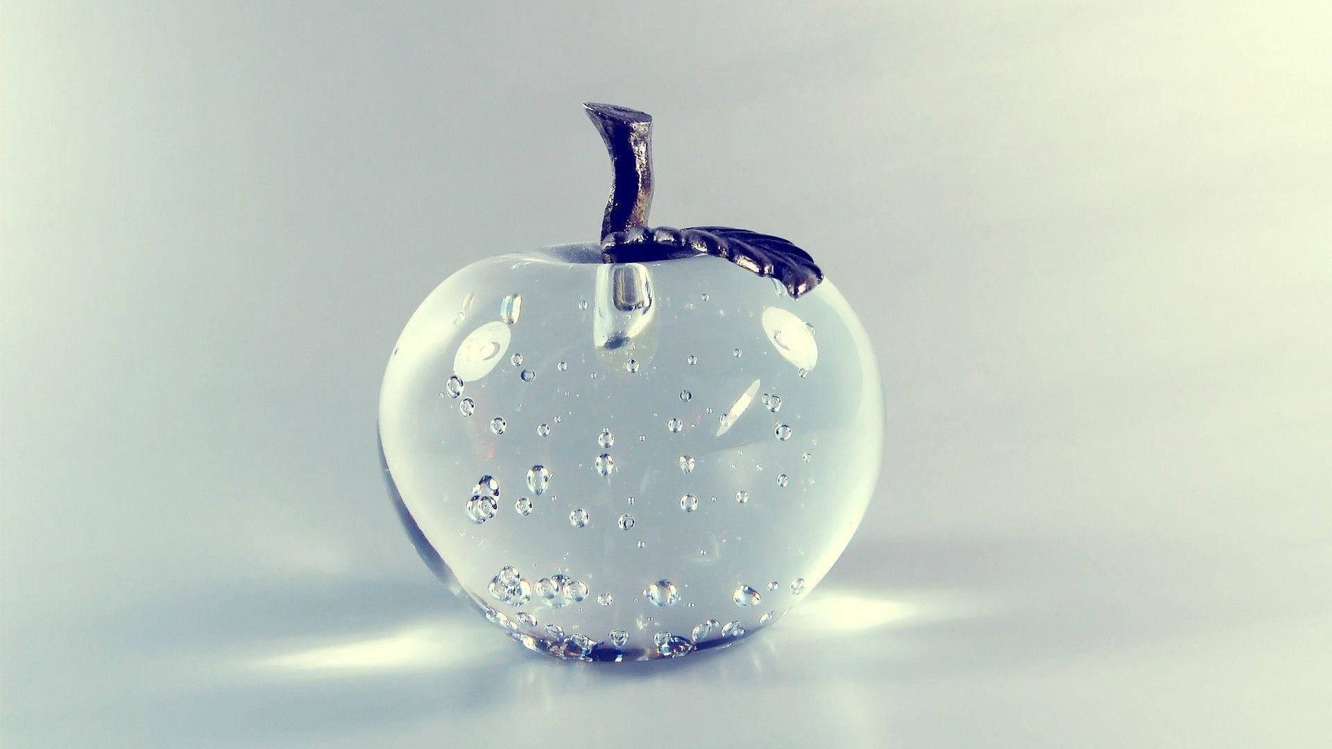 水晶玻璃蘋果 壁紙 — 一個透明玻璃蘋果雕塑，內嵌氣泡，帶有深色莖部，放置在反光表面上，背景為柔和的灰色。