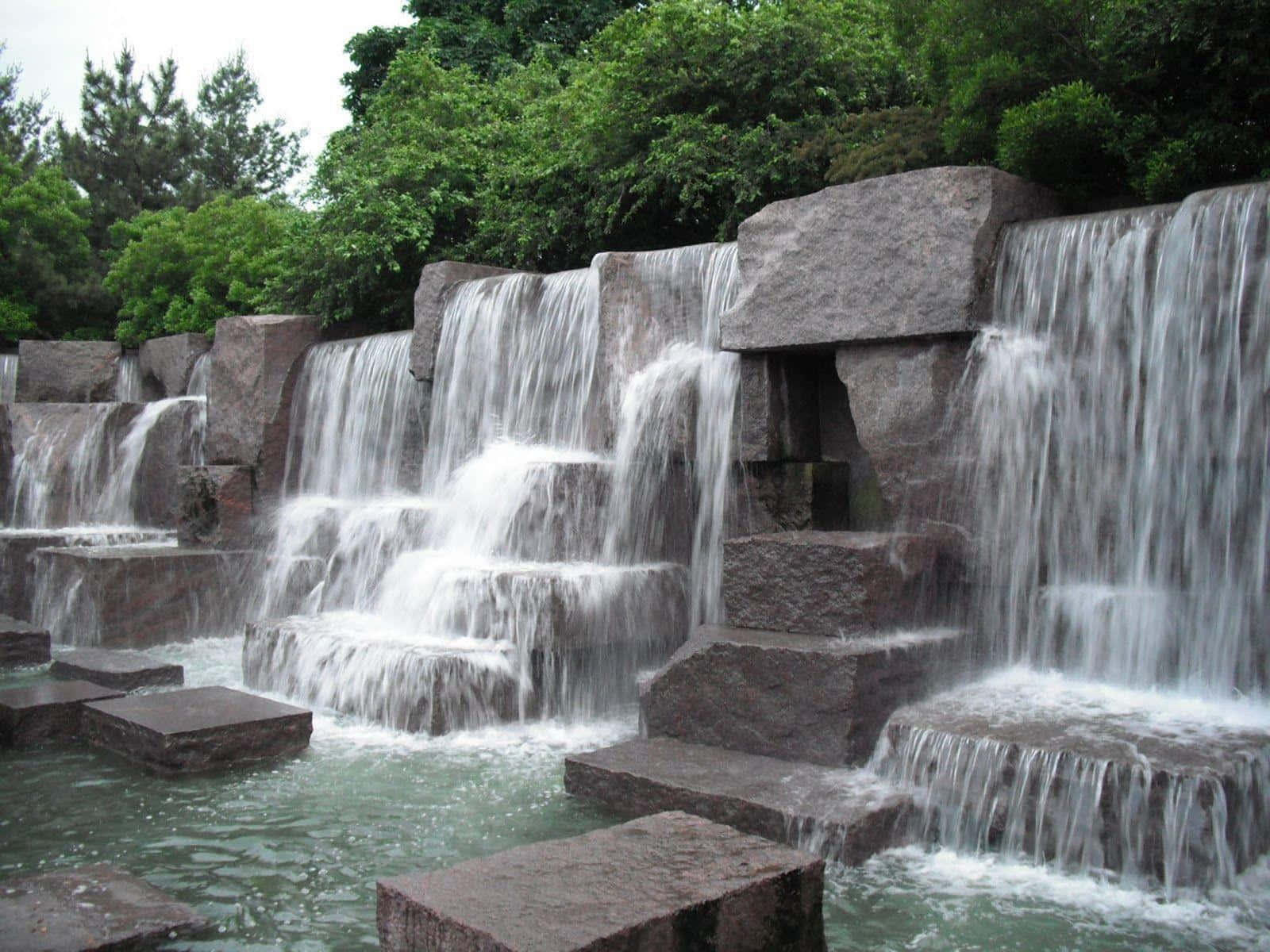 Health And Beauty 桌布 壁紙 — 水流優雅地從層疊的深色石塊上傾瀉而下，流入一個被茂密綠樹環繞的寧靜水池。