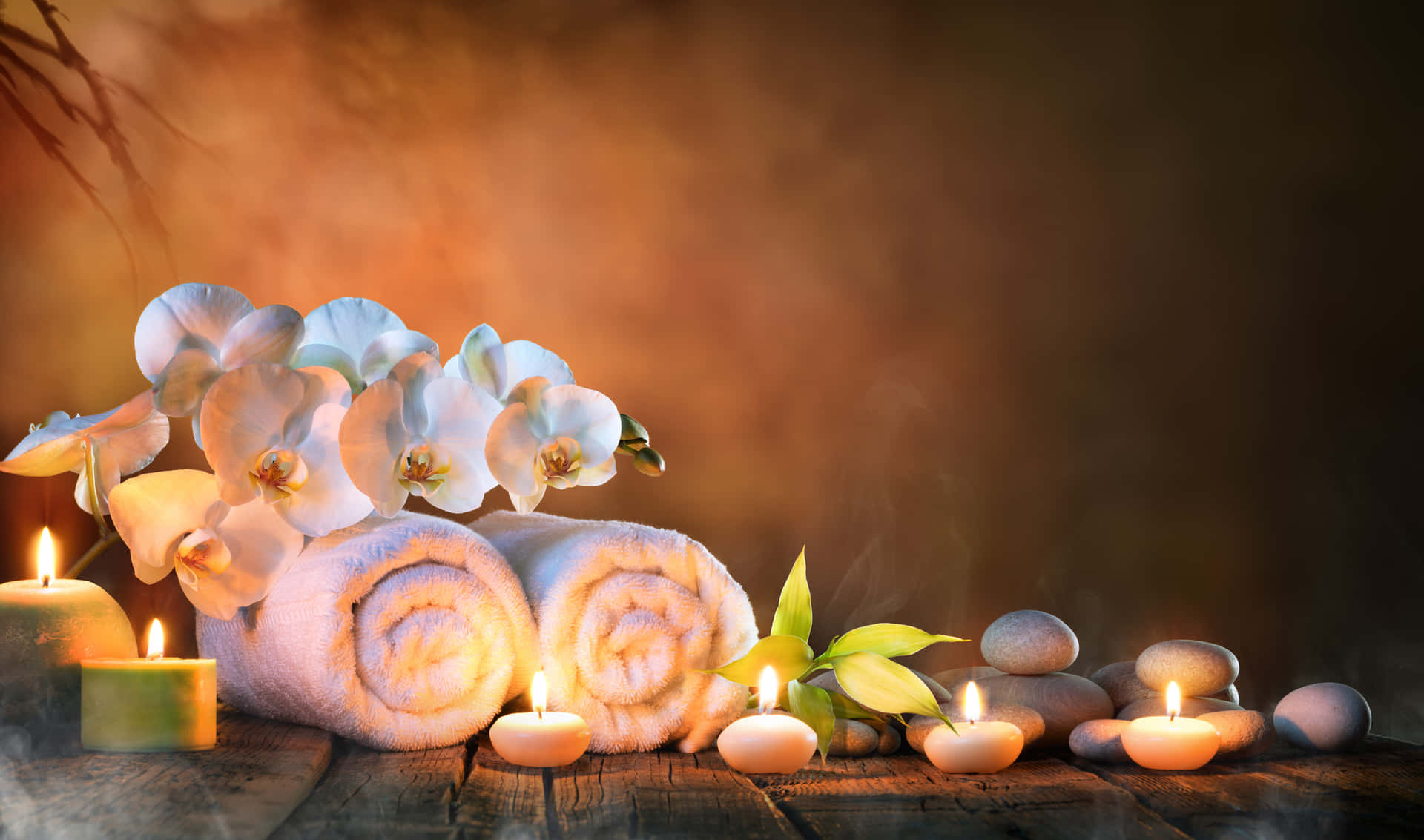 水療背景：白色蘭花、白毛巾與蠟燭 壁紙 — 柔和光線的水療場景，在質樸的木頭上擺放著捲起的毛巾、蘭花、蠟燭和石頭，營造寧靜氛圍。