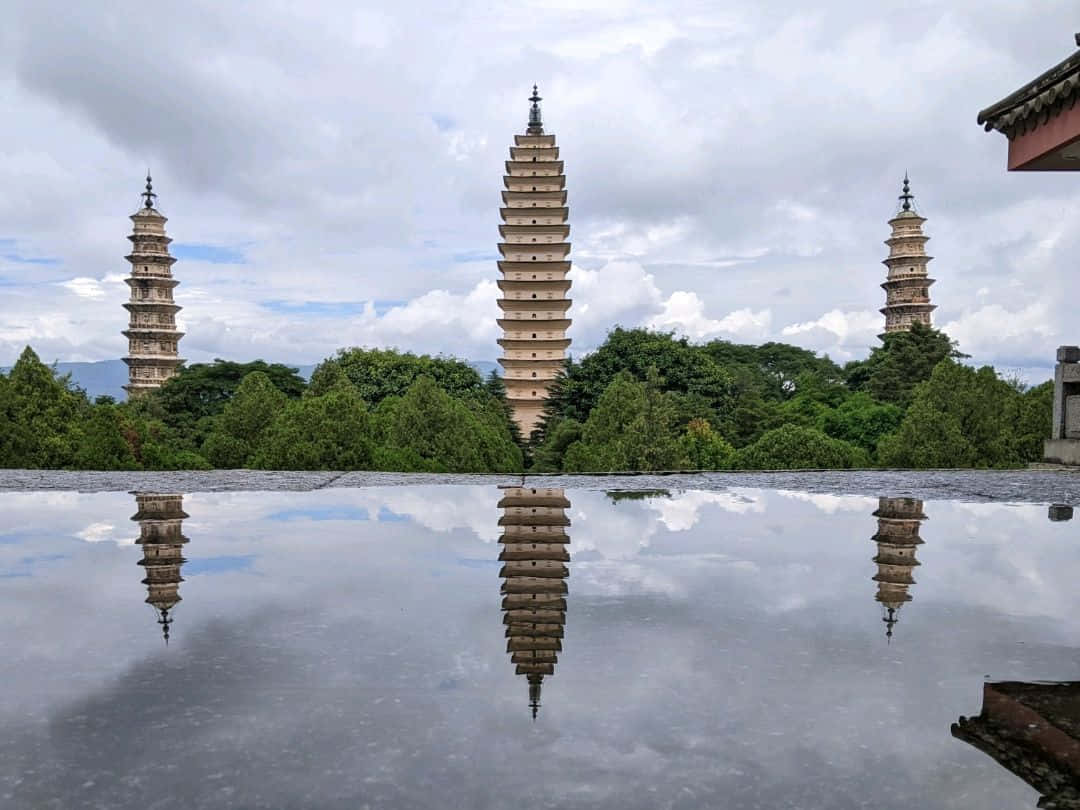 水面倒影三塔 壁紙 — 三座古老石塔矗立在茂密綠樹後方，完美倒映在平靜的池塘中，天空多雲。