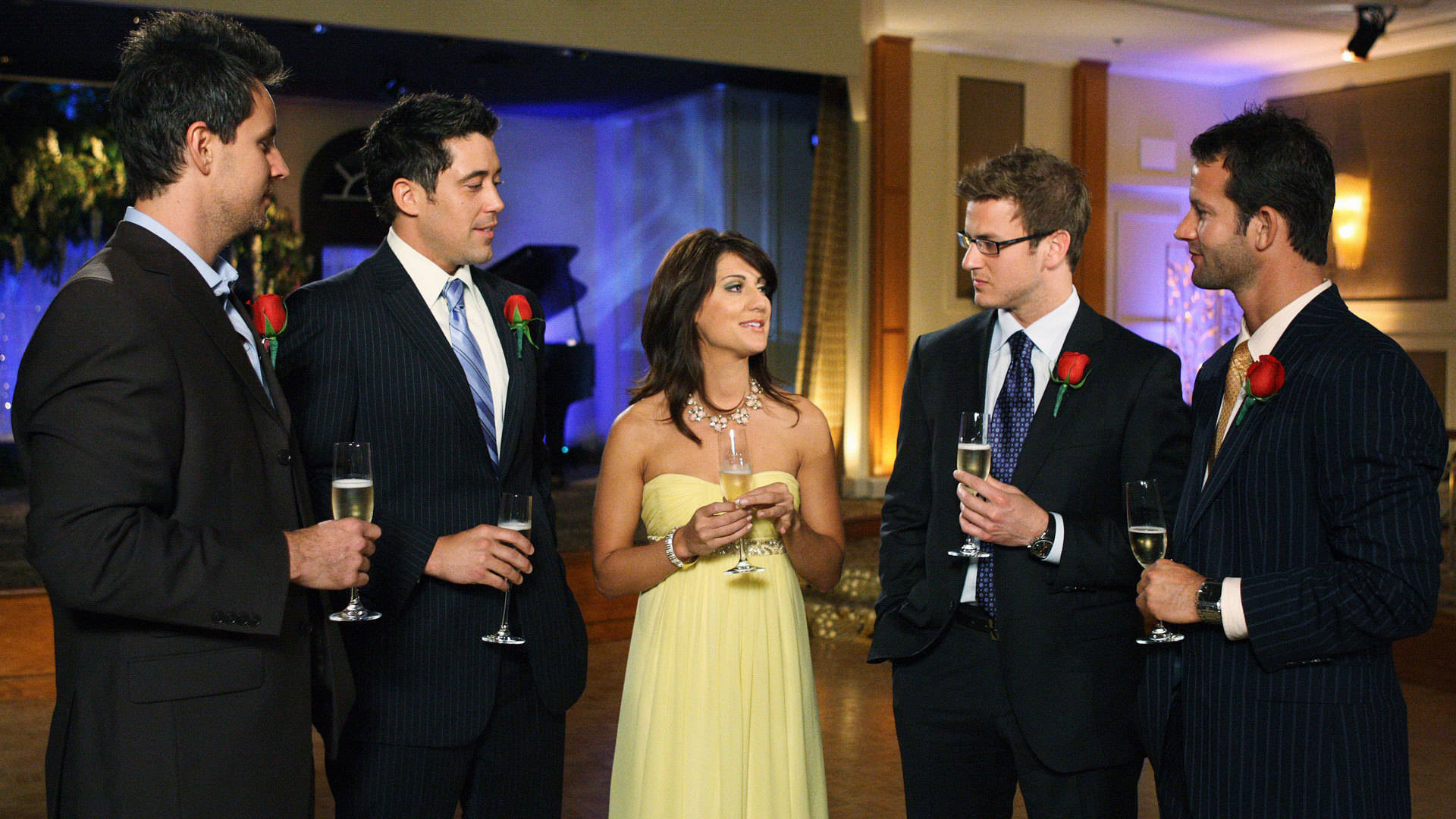 《求愛大作戰》吉莉安·哈里斯與眾男士 壁紙 — 五位穿著正式服裝的賓客在優雅的室內派對中交談，手持香檳杯和紅玫瑰。