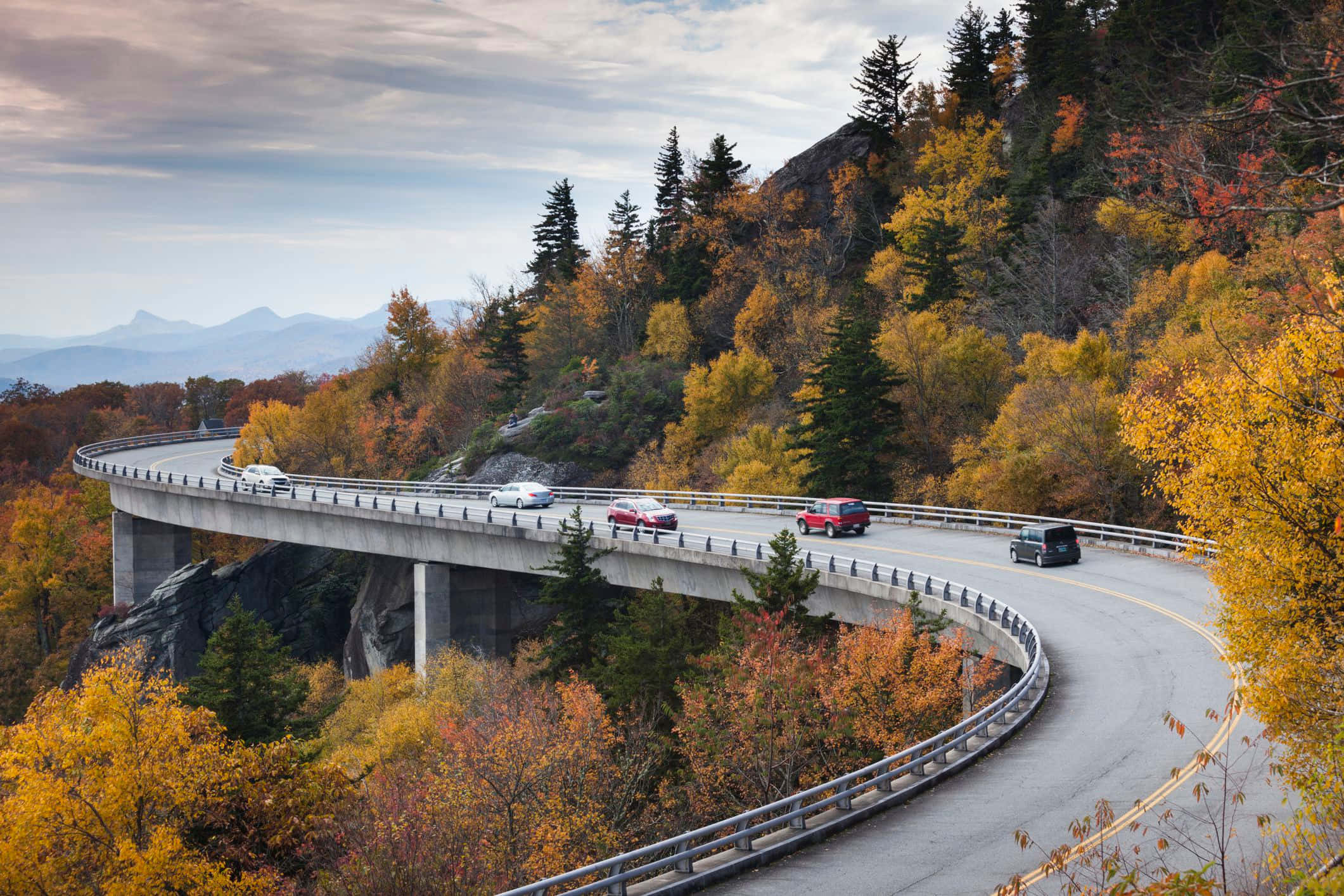 Blue Ridge Parkway 桌布 壁紙 — 汽車在蜿蜒的山區高速公路上行駛，周圍環繞著鮮豔的秋葉，天空多雲。