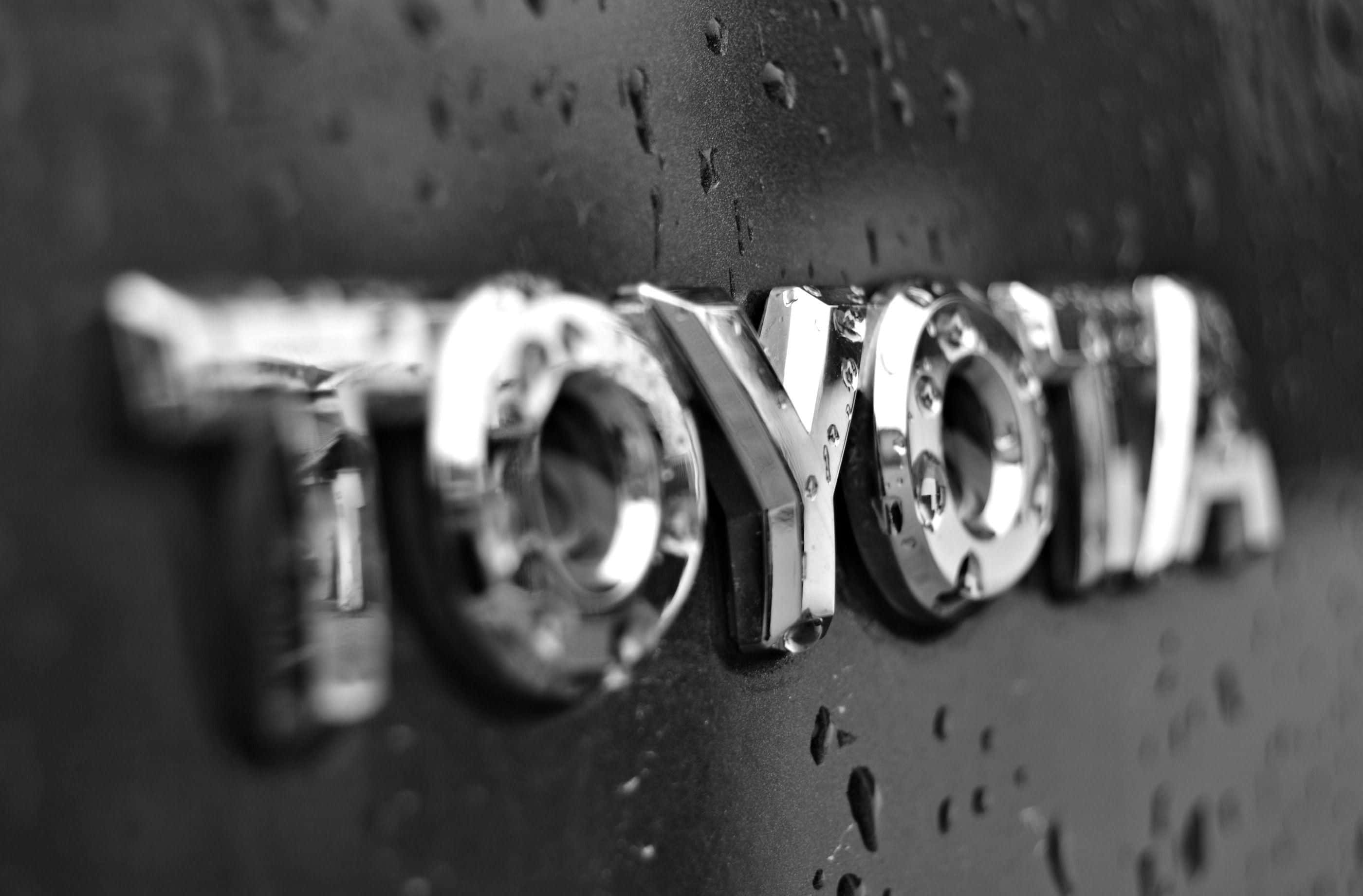 Toyota 桌布 壁紙 — 汽車引擎蓋上濕透的 Toyota 標誌特寫，雨滴在單色調中閃爍。