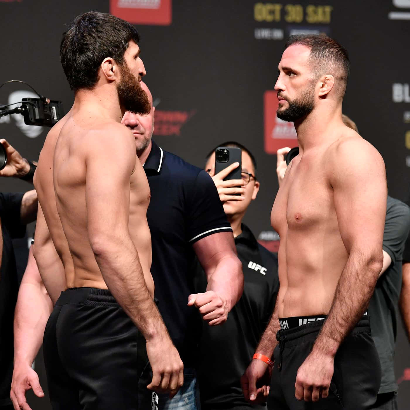 兩位赤裸上身的MMA拳手在UFC稱重活動中互相凝視，周圍有攝影師。