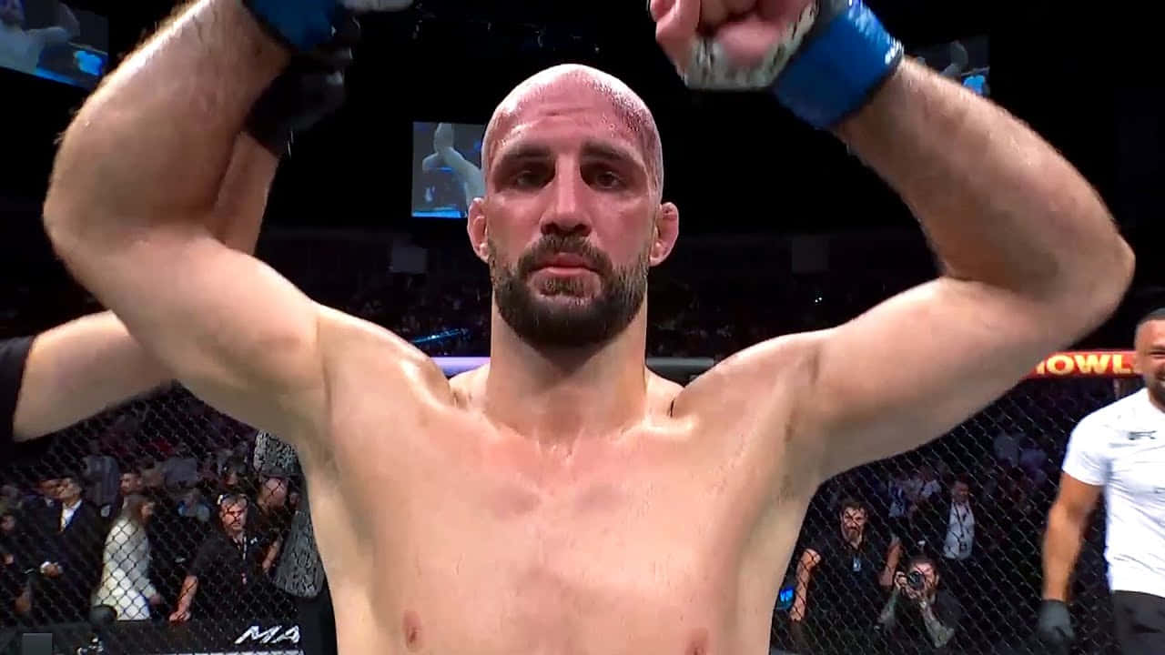 一位光頭鬍鬚的MMA拳手在八角籠內高舉拳頭慶祝勝利。