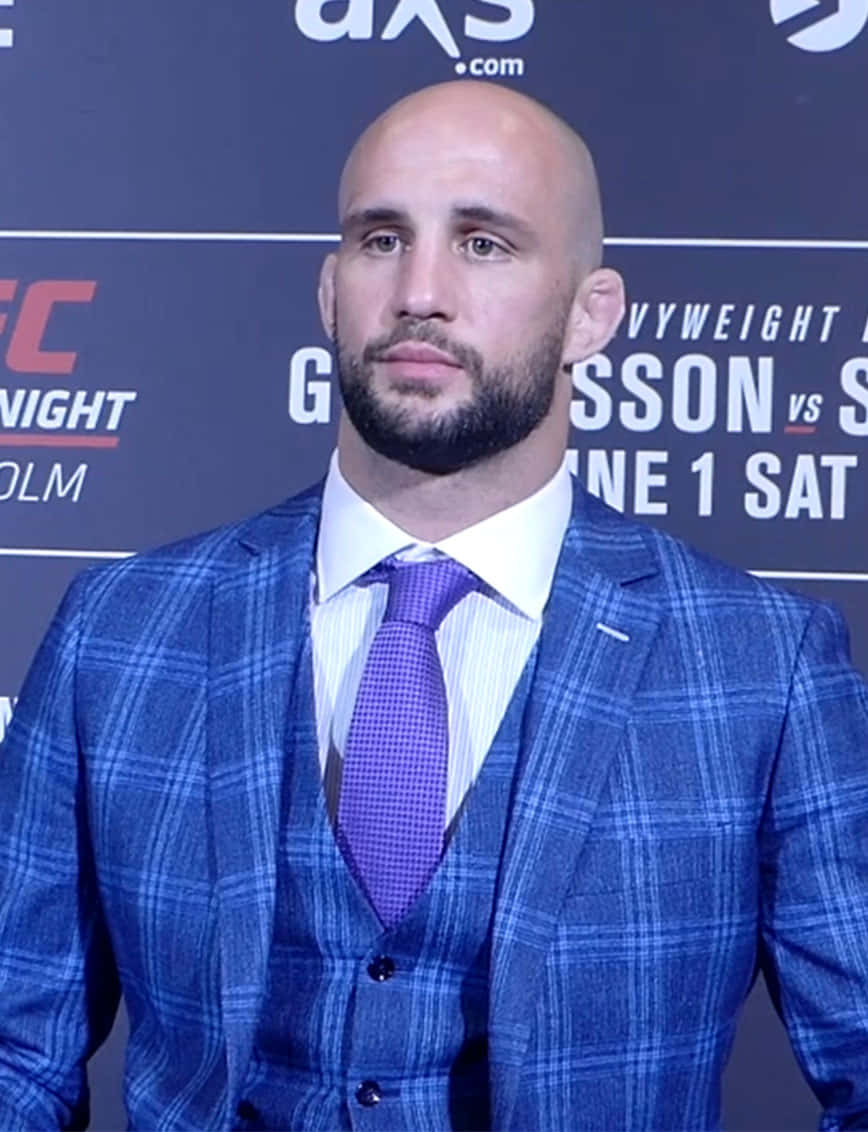 禿頭男子在UFC記者會上穿著藍色格紋西裝和紫色領帶。