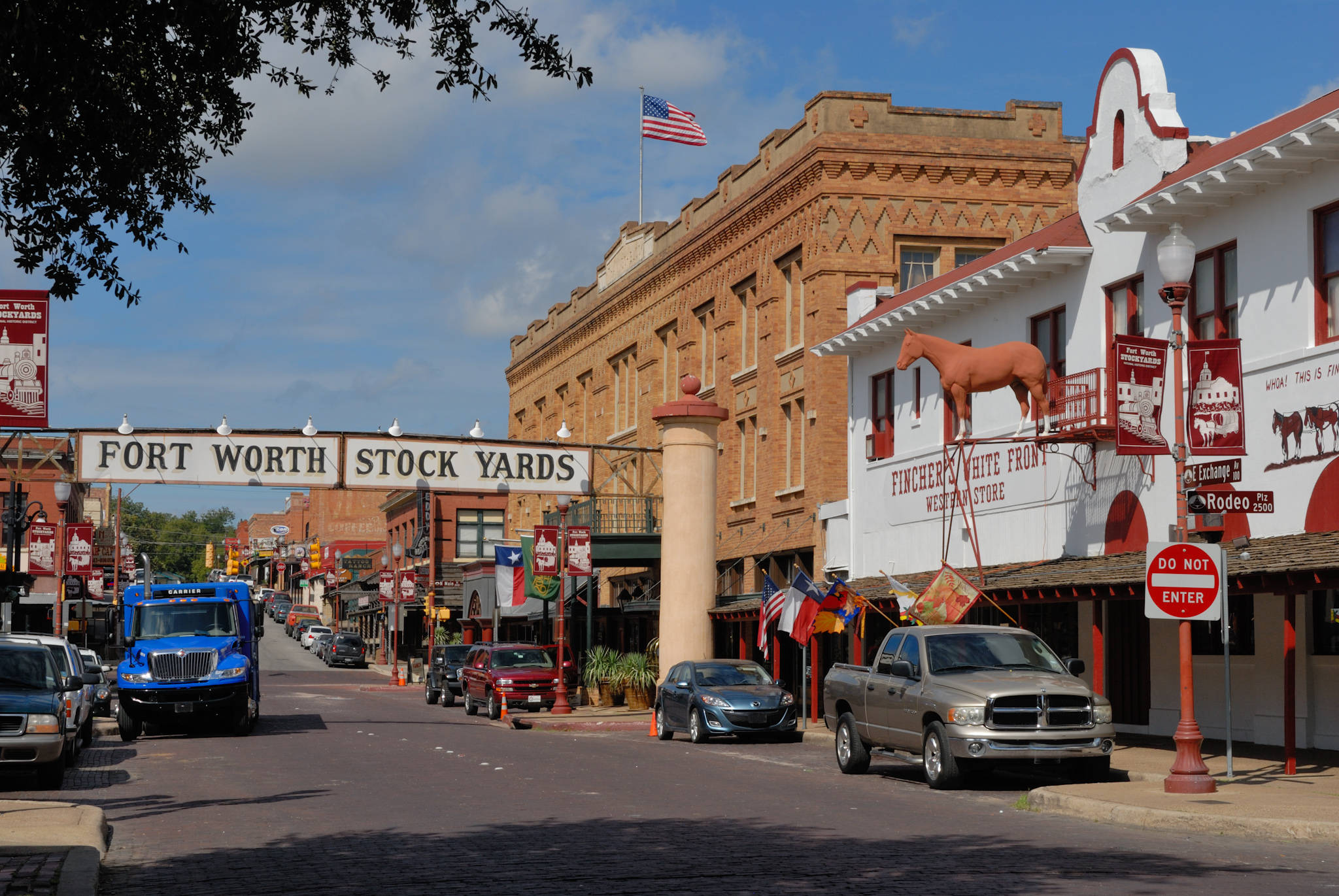 沃斯堡牲畜市場 壁紙 — Fort Worth Stock Yards 一條陽光普照的街道，有歷史悠久的磚砌建築、馬匹雕像和一輛藍色貨車停泊在晴朗的藍天下。