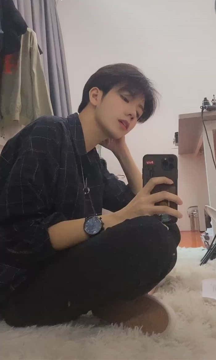 沉思人生的美學男孩 壁紙 — 一位身穿深色格仔衫嘅年輕男子交叉腳坐喺柔軟嘅白色地氈上，喺燈光柔和嘅睡房入面影緊鏡自拍。