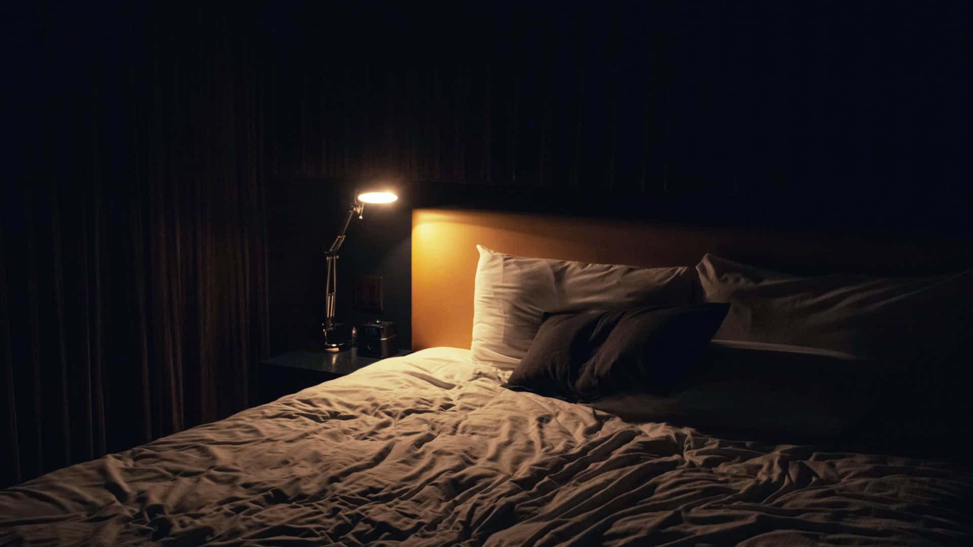 沉浸於夜間的美麗 壁紙 — 夜晚昏暗的臥室，一盞燈在凌亂的床和深色窗簾上投下溫暖的光芒。