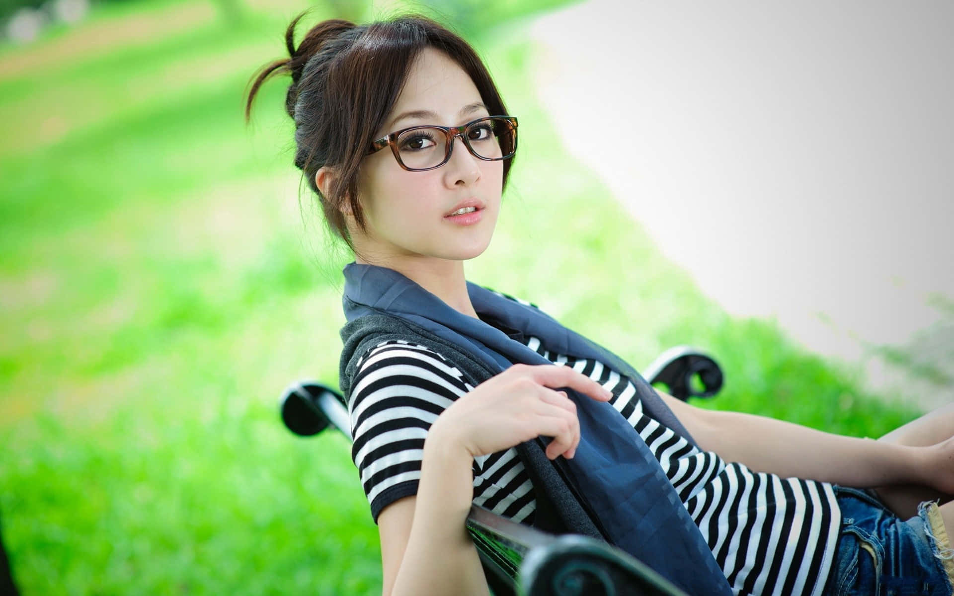 沉浸於大自然之美 壁紙 — 一位戴眼鏡、身穿條紋襯衫的年輕女性，在充滿活力的綠色草地背景襯托下，悠閒地躺在公園長椅上。