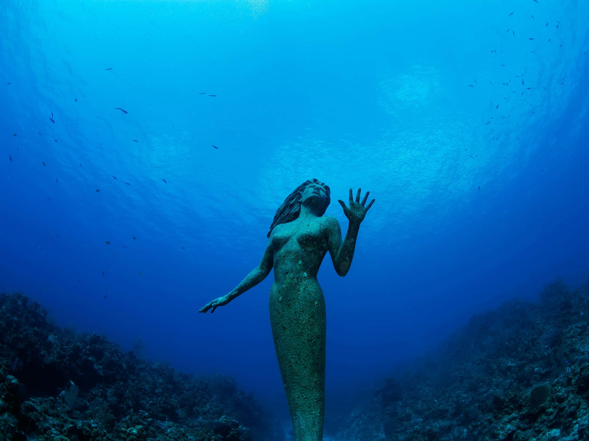 沉浸於海底世界的美麗與壯麗 壁紙 — 一尊青銅美人魚雕像在水中優雅地張開雙臂站立，陽光穿過上方深藍色的海洋照射下來。