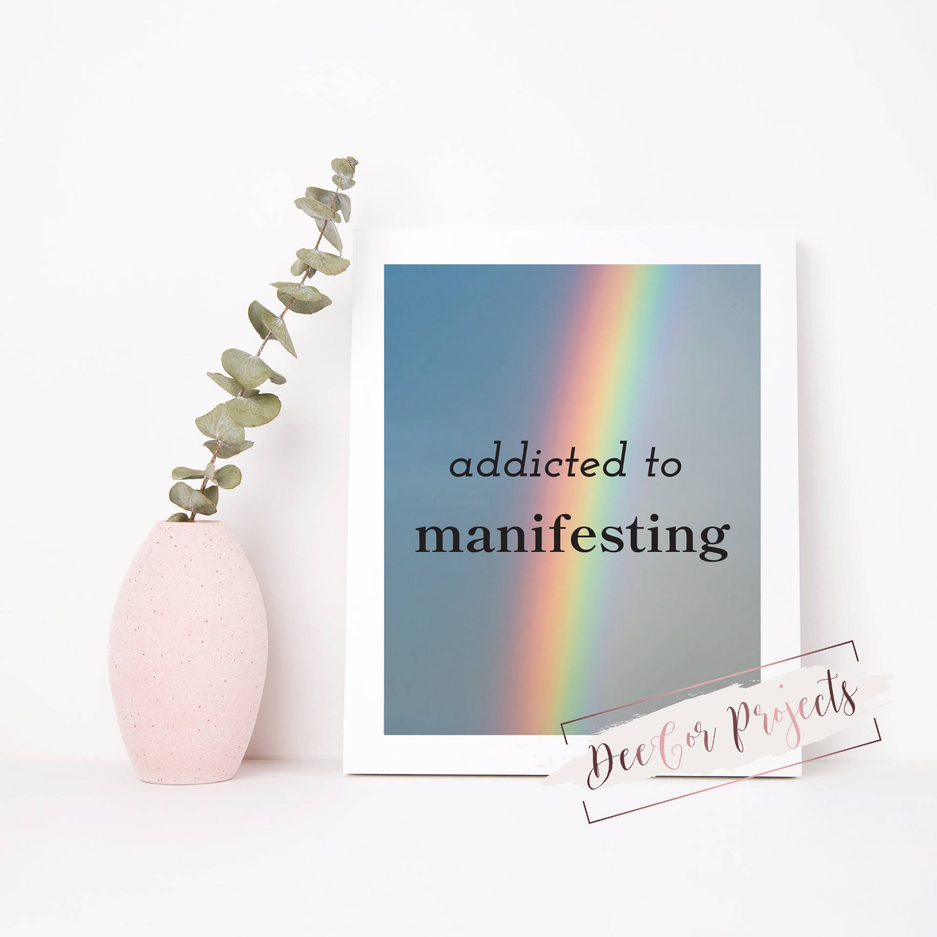 裱框印刷品，帶有彩虹和文字「addicted to manifesting」，旁邊是一個粉紅色花瓶，插著尤加利葉。