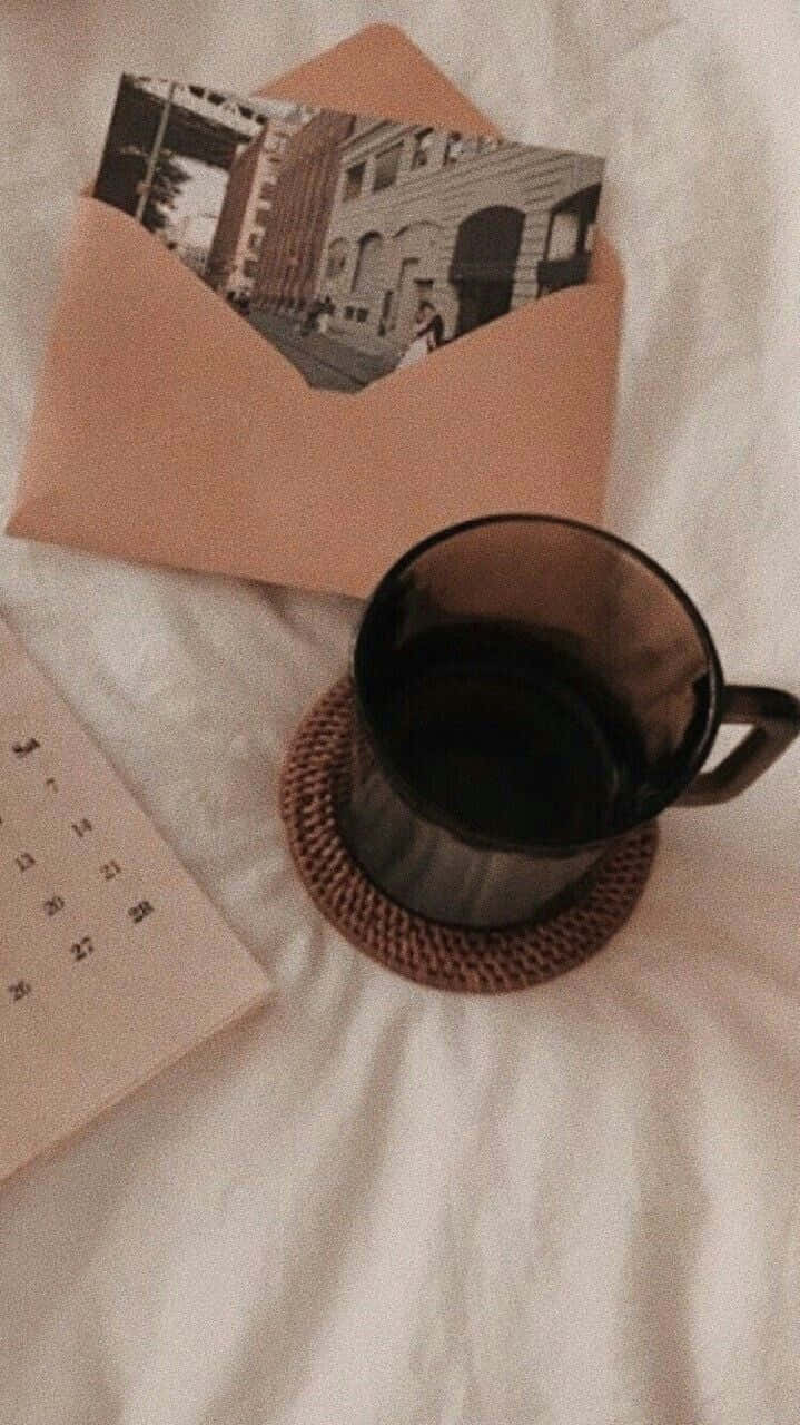 沉醉於棕色美學的時尚魅力 壁紙 — 一個黑色馬克杯放在編織杯墊上，旁邊有一個桃色信封，內含一張復古街頭照片和一本筆記本，置於柔軟的白色布料上。