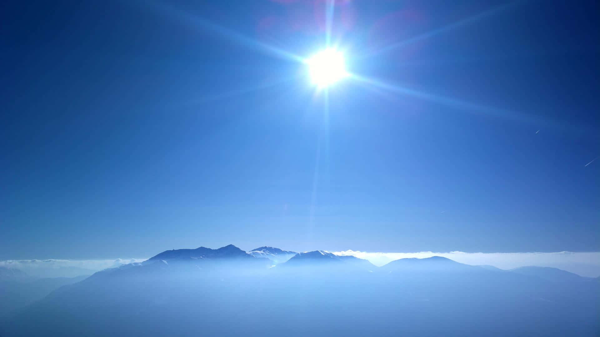 沉醉於美麗藍天 壁紙 — 在廣闊清澈的藍天下，一輪燦爛的太陽散發出強烈的光芒，照耀著被大氣薄霧籠罩的遙遠山脈。