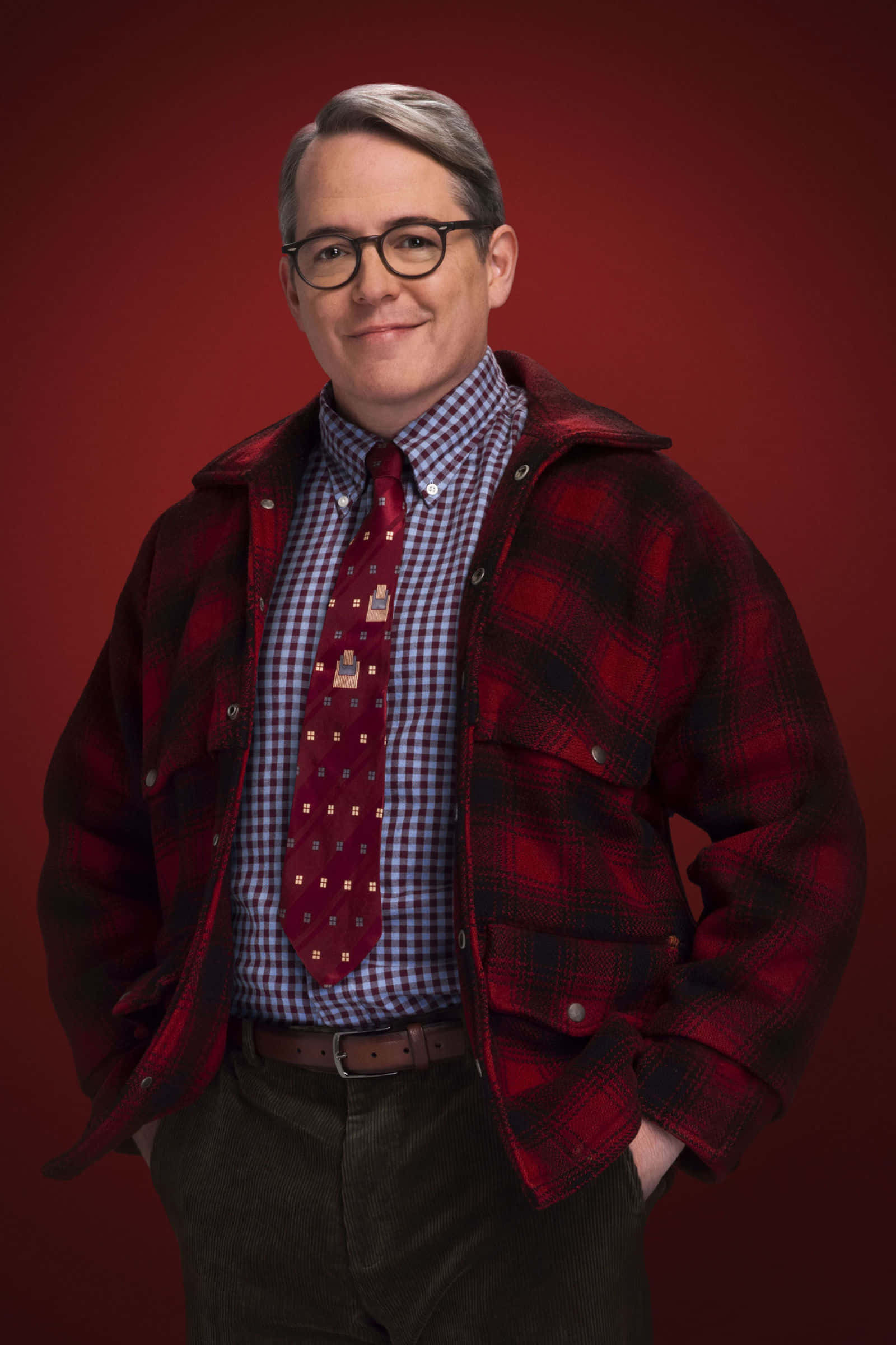 沒有什麼比《A Christmas Story》更能傳遞聖誕的溫暖 壁紙 — 一名戴眼鏡的男子，身穿紅色格仔外套，內搭格仔恤衫和圖案領帶，在深紅色背景前溫暖地微笑。
