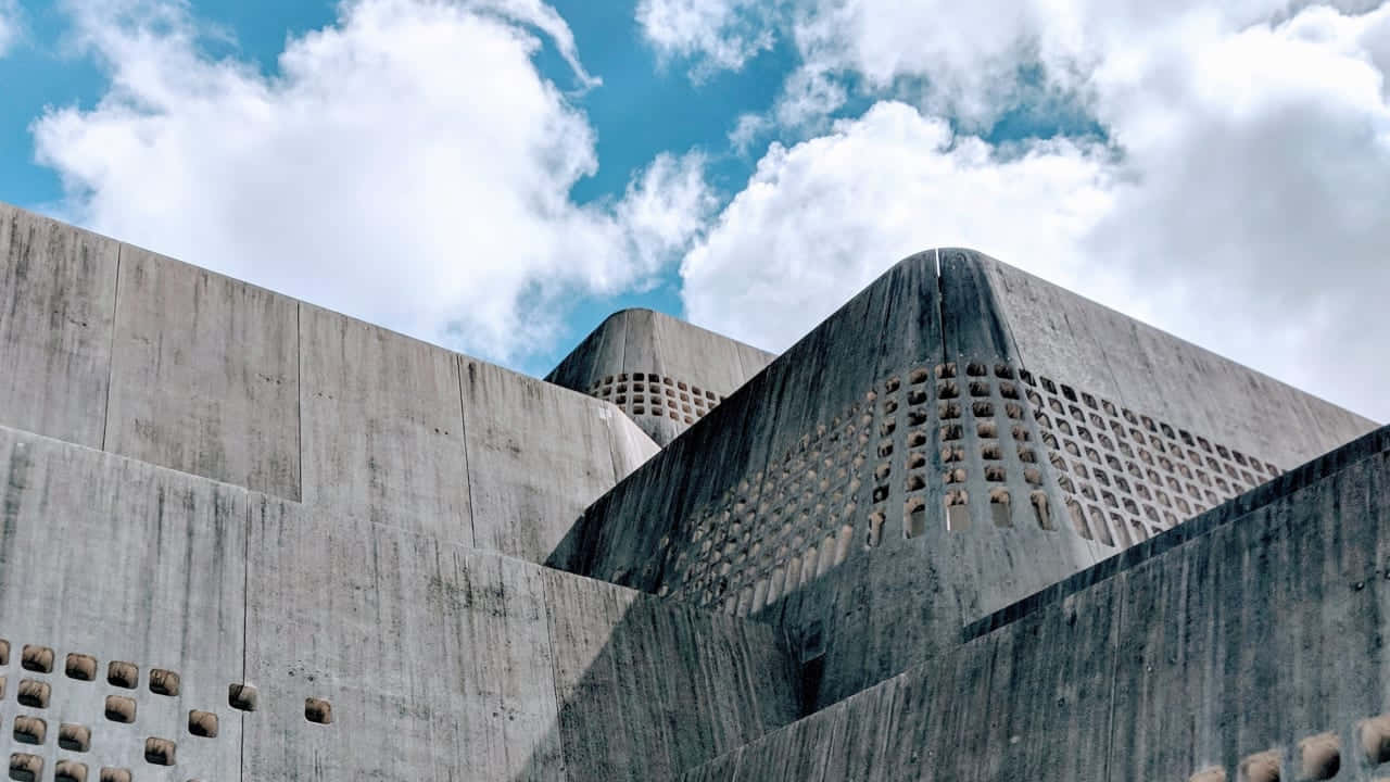 沖繩混凝土博物館圖片 壁紙 — 低角度拍攝的粗獷主義混凝土建築，具有幾何形狀和穿孔牆壁，背景是明亮的藍天和蓬鬆的白雲。