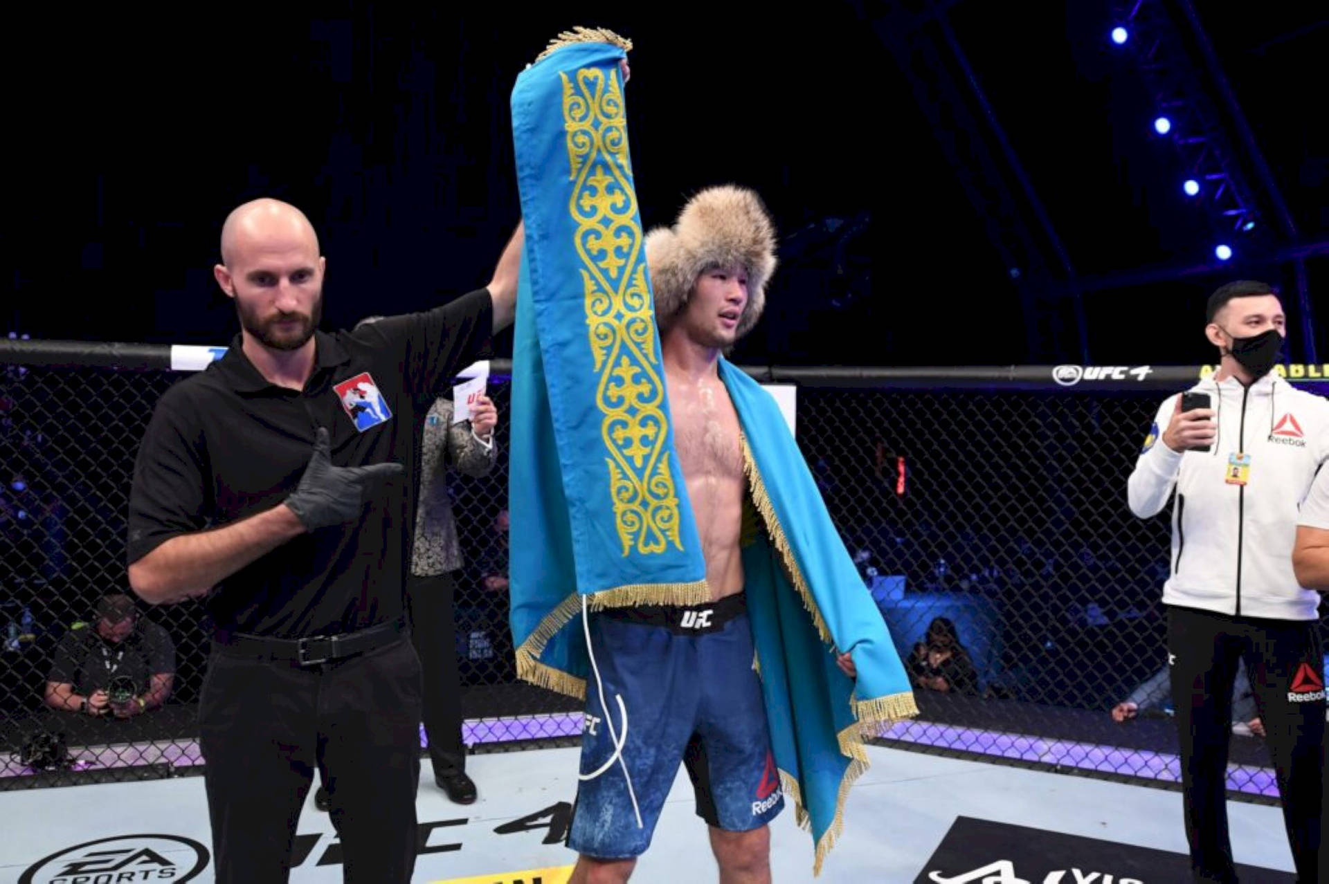 沙夫卡特·拉赫莫諾夫在八角籠內舉起哈薩克國旗 壁紙 — 一位勝利的UFC拳手披著藍金色華麗的哈薩克旗幟，站在八角籠中，裁判舉起他的手。