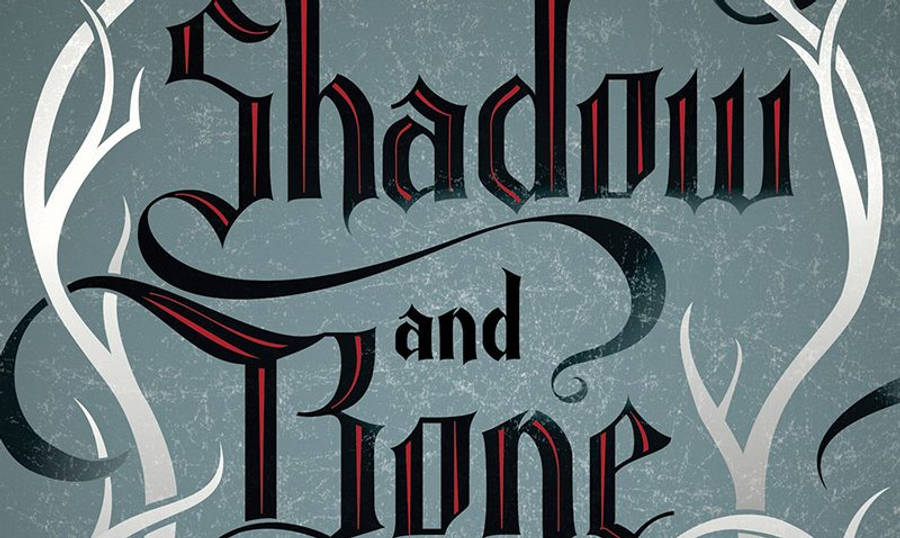 《沙影與骨》書籍封面 壁紙 — 優雅的哥德式字體拼寫 Shadow and Bone，紅黑字體在帶紋理的灰藍色背景上，由白色華麗漩渦框住。