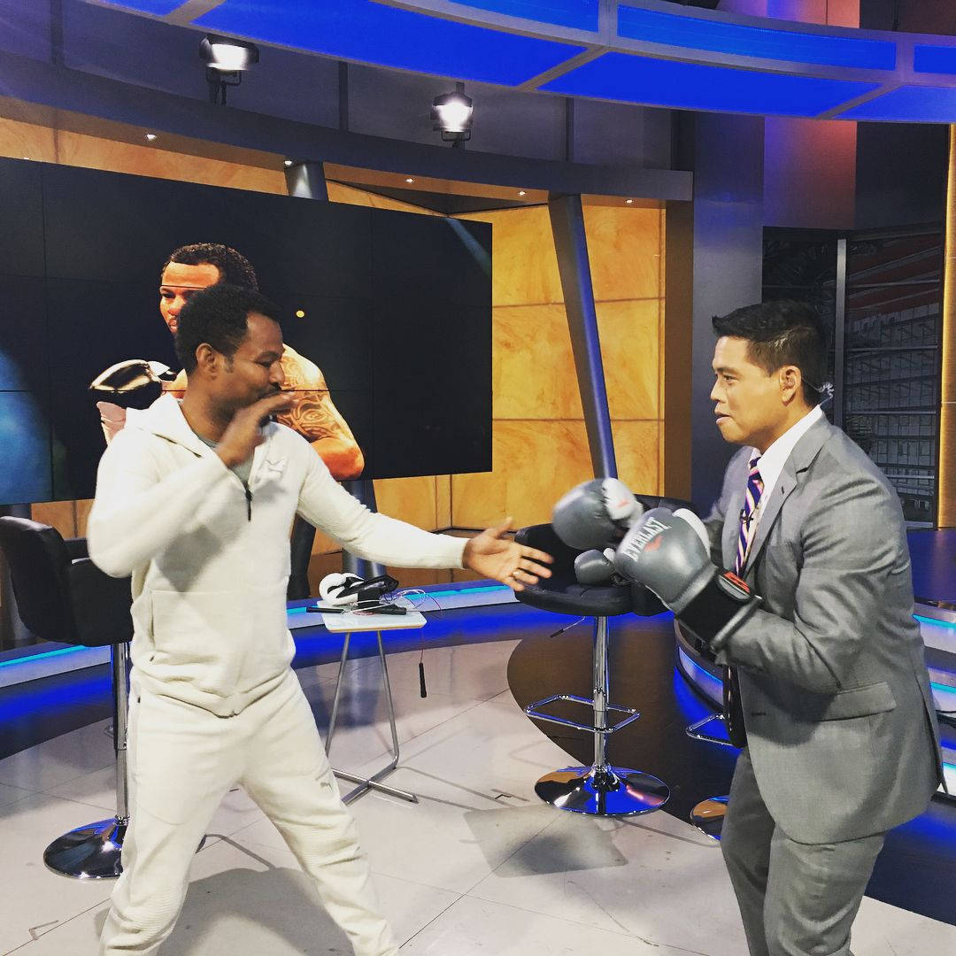 沙恩·莫斯利教電視主持人出拳 壁紙 — 兩名戴著拳擊手套的男子在一個現代電視錄影廠內對練，現場有藍色燈光和木質牆板。