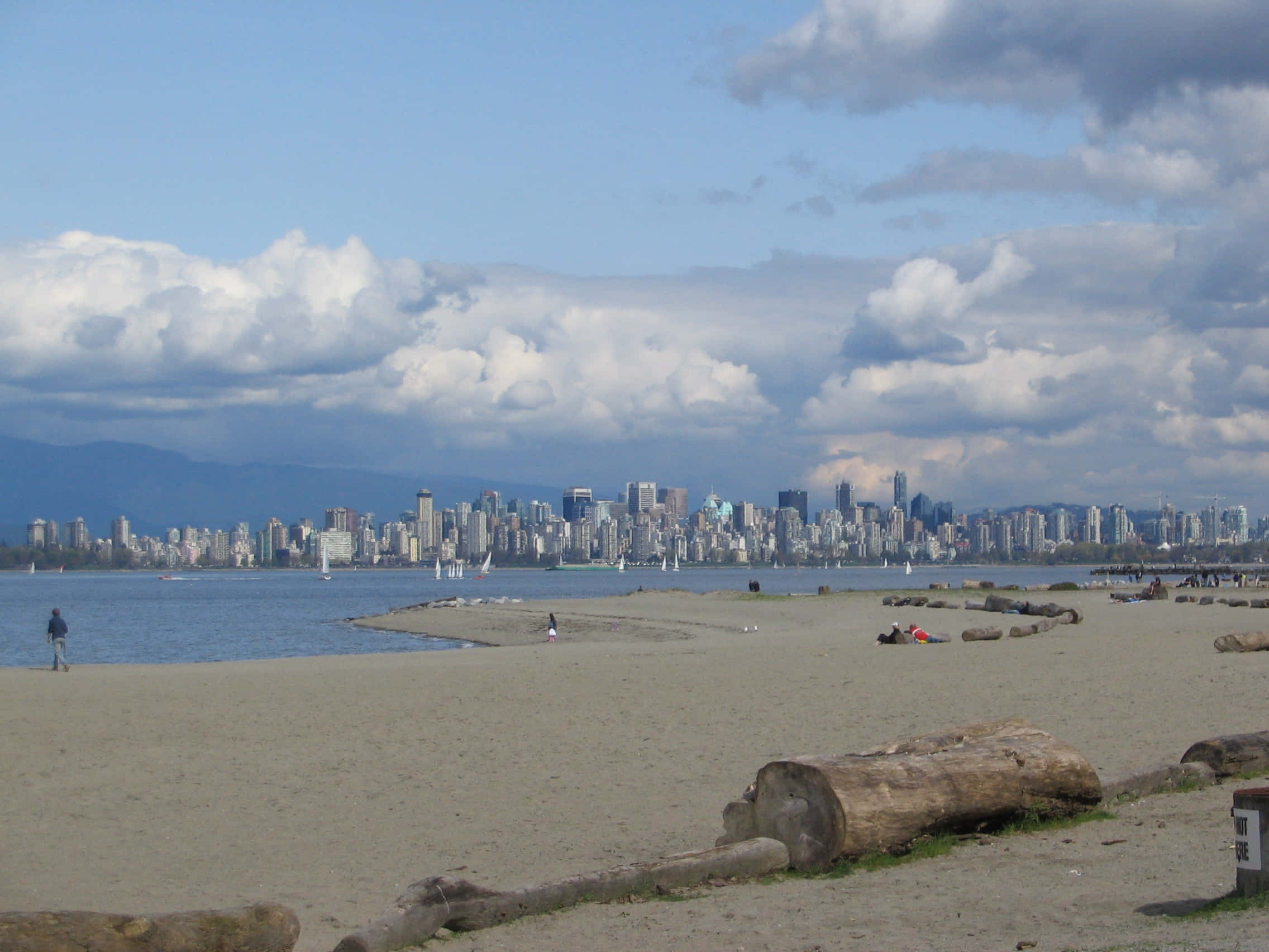 Spanish Beach 桌布 壁紙 — 沙灘前景有漂流木，越過水面可看到城市天際線，天空多雲。