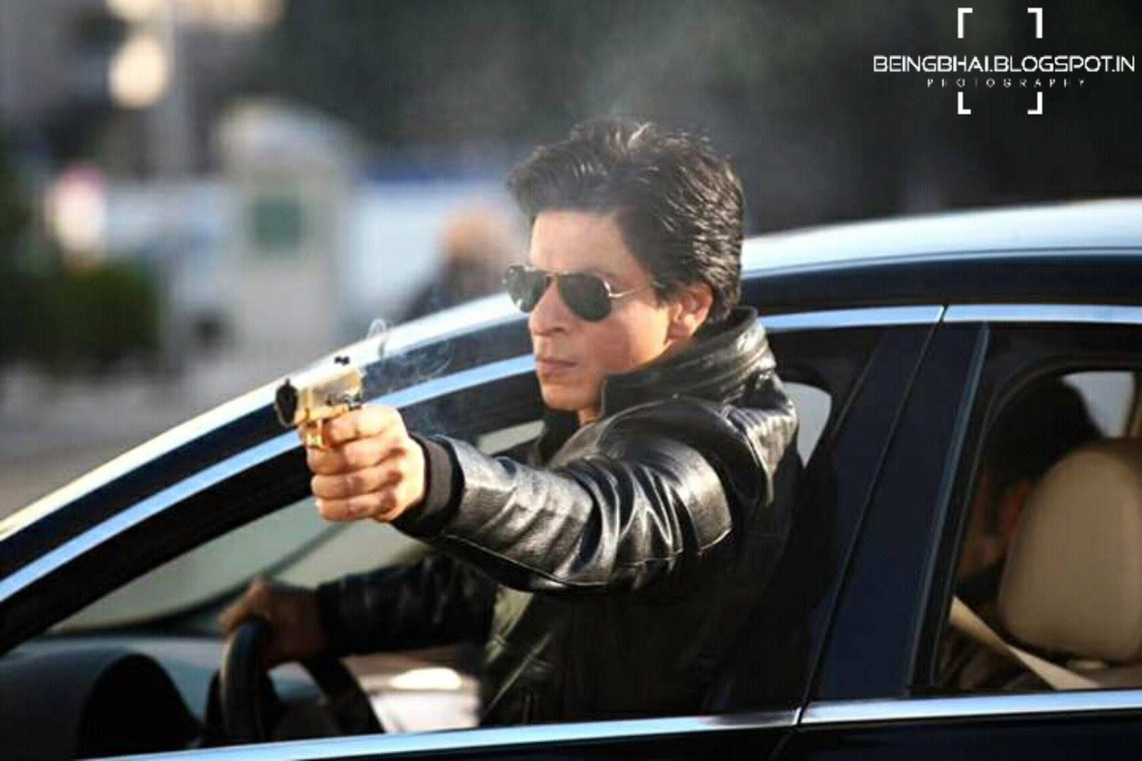 沙魯克·汗高清《Dilwale》劇照 壁紙 — 一名身穿黑色皮褸和太陽眼鏡的男子在日間從車窗開槍，手槍冒煙。